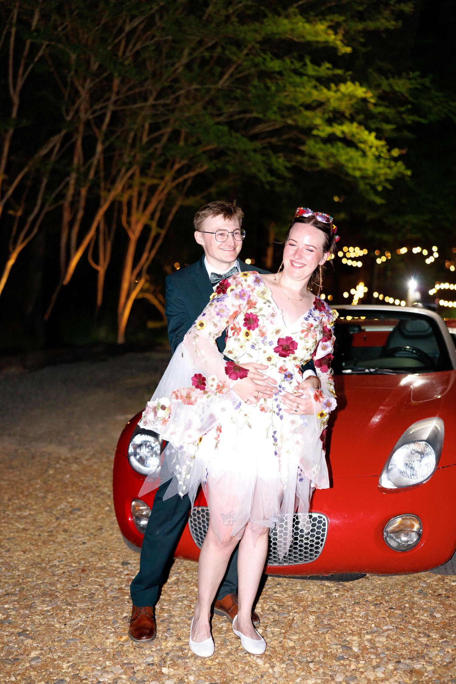 Couple posing in front of a red sports car at night, woman in floral dress with tulle skirt, man in dark suit.