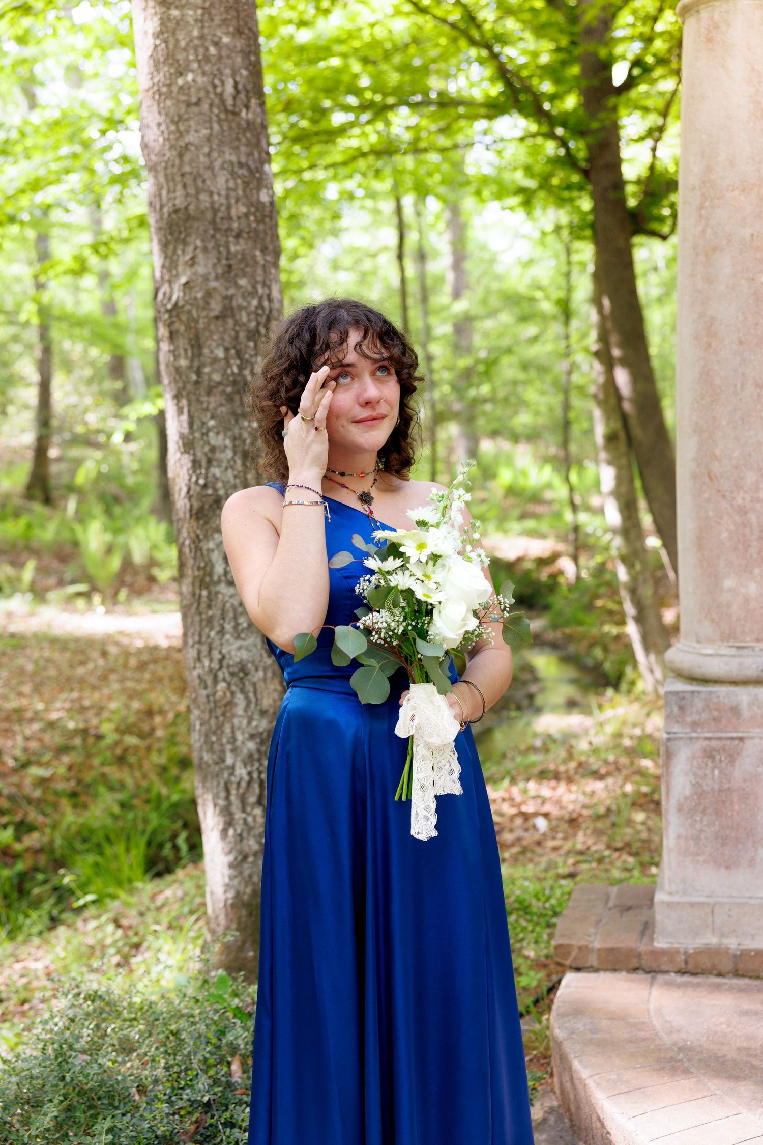 Bridesmaid in blue gown wiping tear holding white bouquet in lush forest wedding setting.