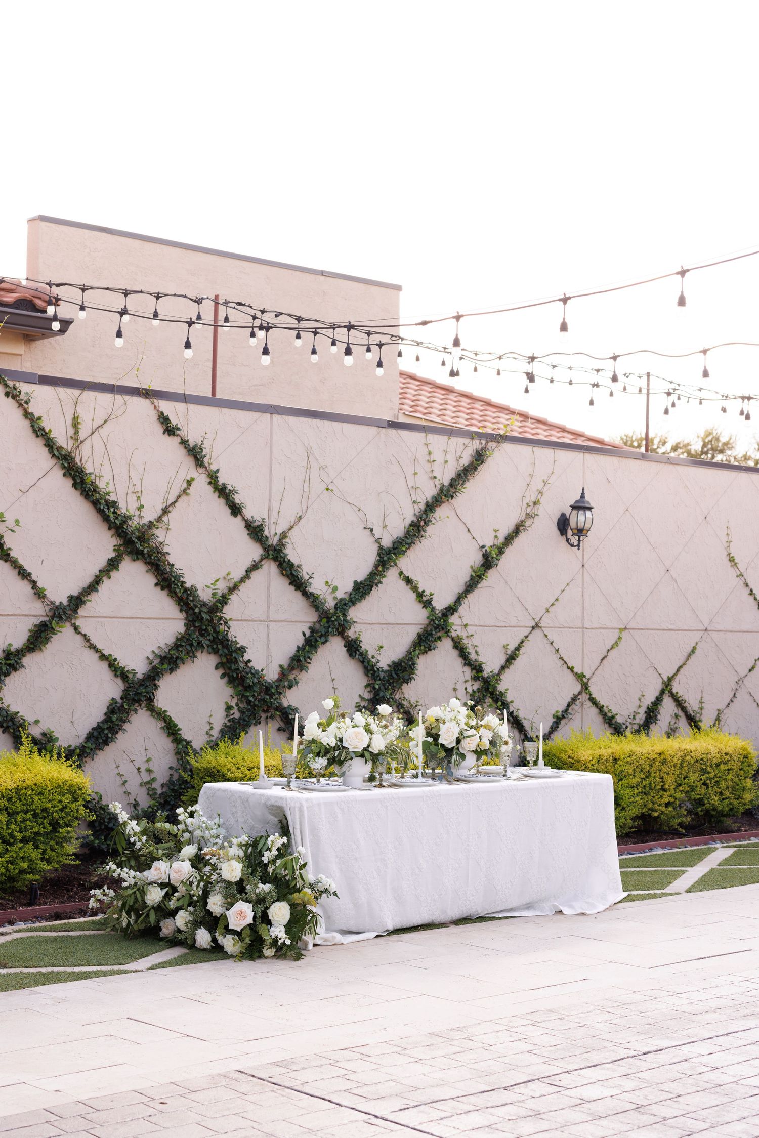 Outdoor wedding reception setup with white tables and climbing vine lattice wall decor.