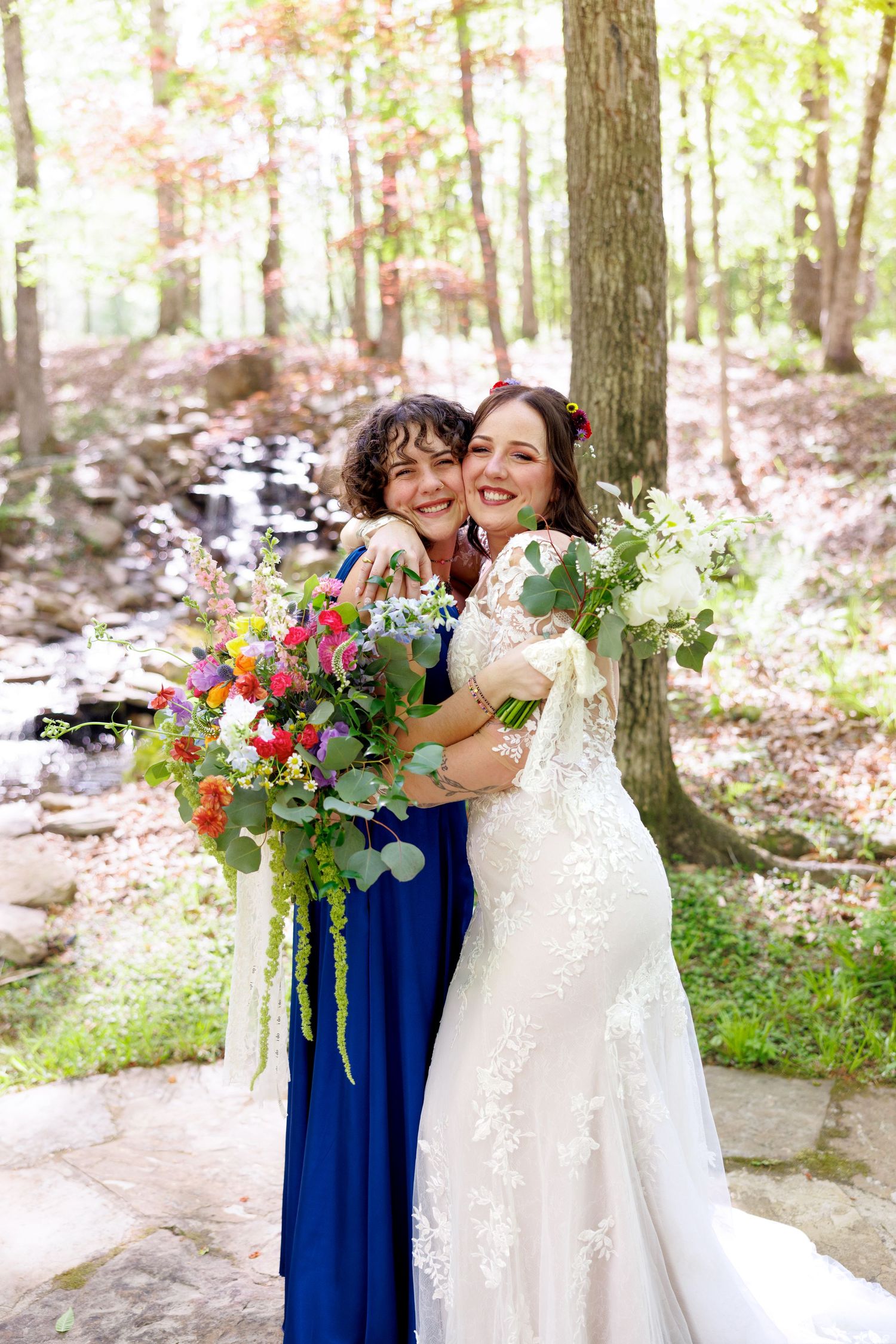 Bride in lace gown and bridesmaid in blue dress hugging outdoors, holding colorful floral bouquets in a woodland setting.