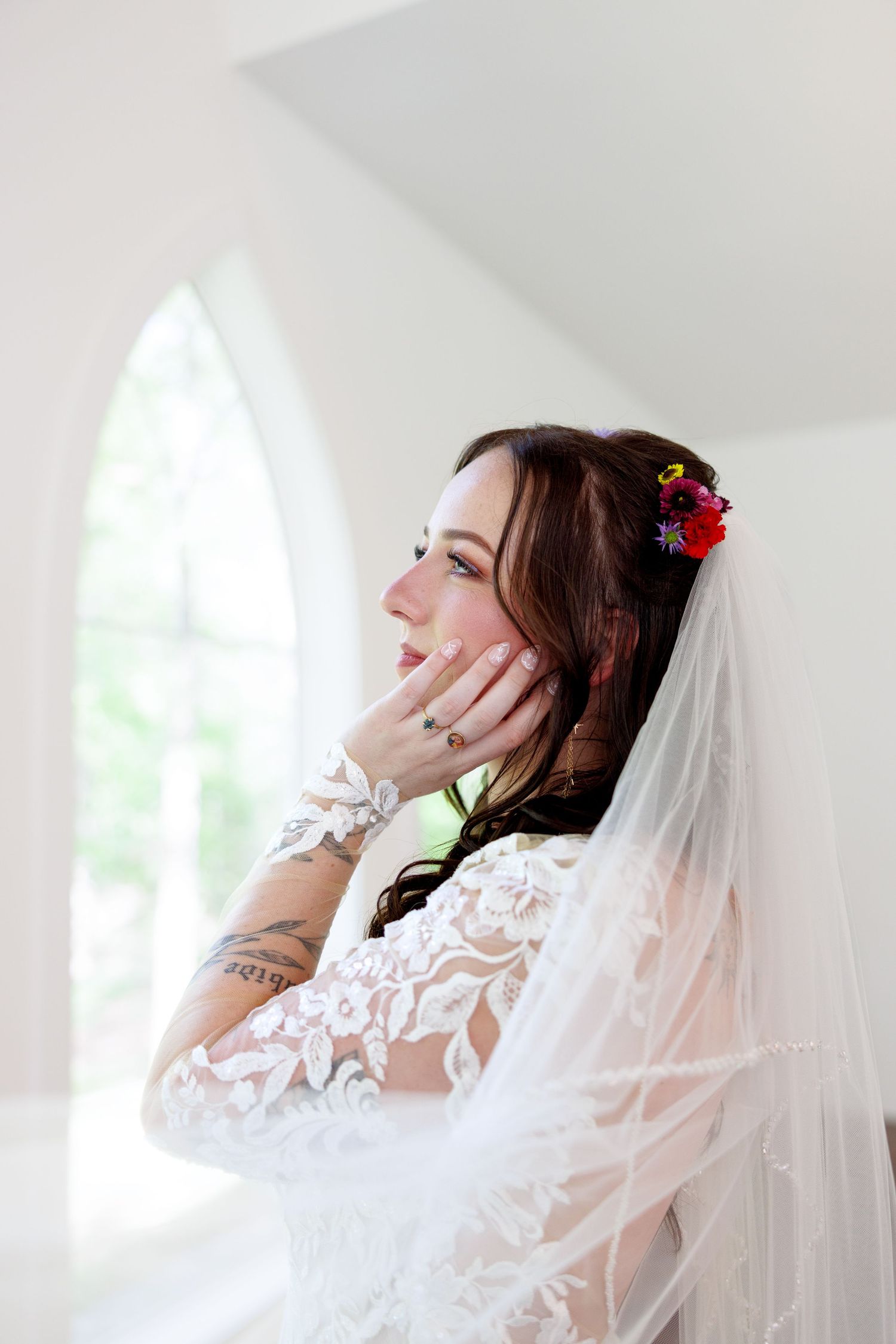 Bride in lace wedding dress and veil with colorful floral hair accessories and tattoos, gazing upward near arched window.