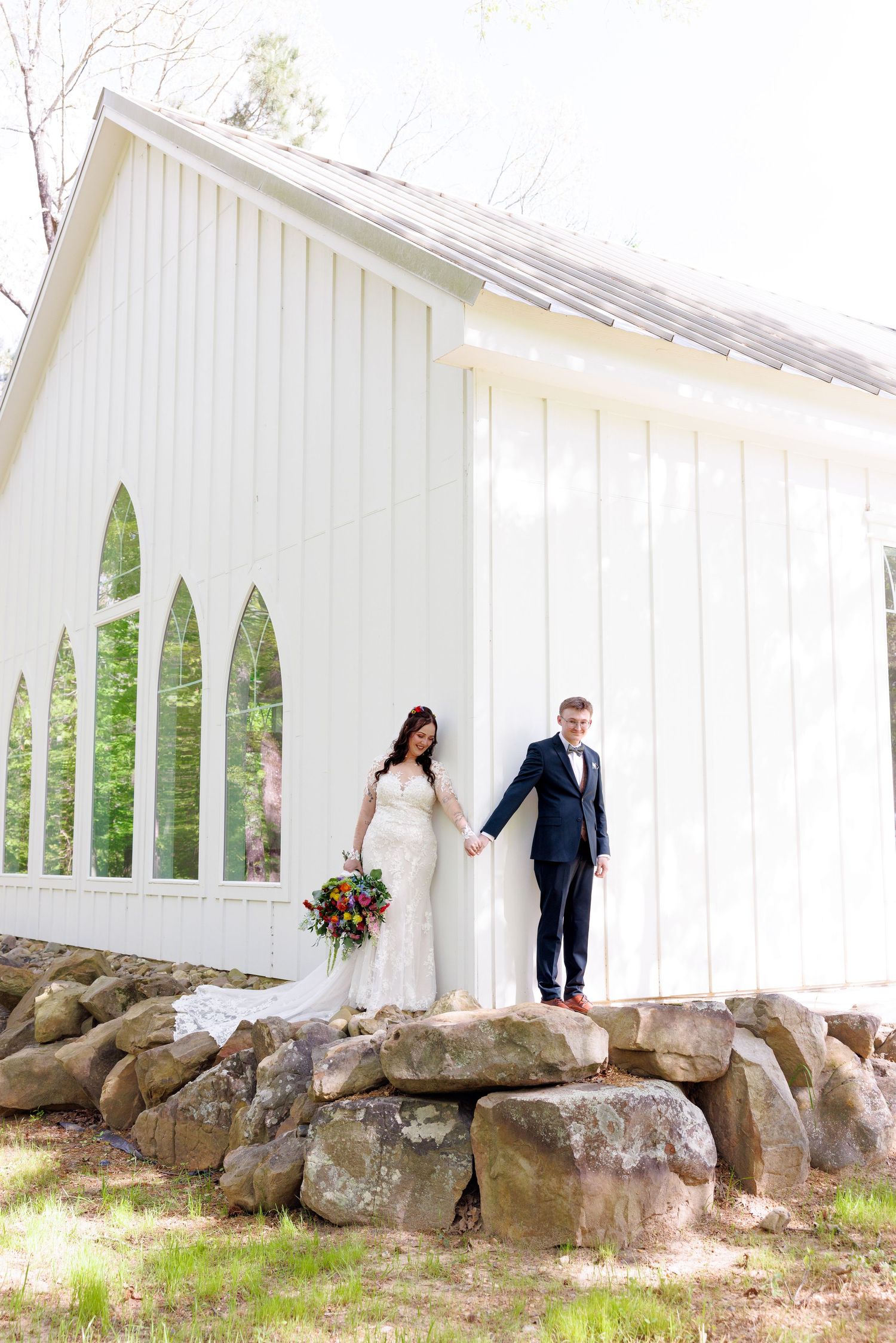 Bride and groom holding hands standing on rocks beside a white chapel with arched windows on their wedding day.