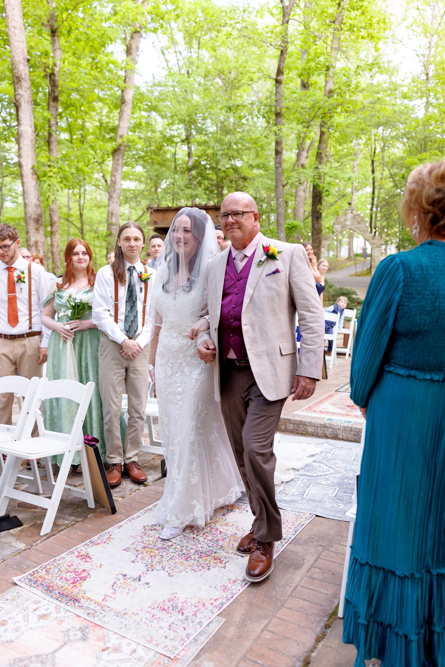 Bride walks down outdoor aisle with escort on colorful rug, guests watching, lush green trees in background.