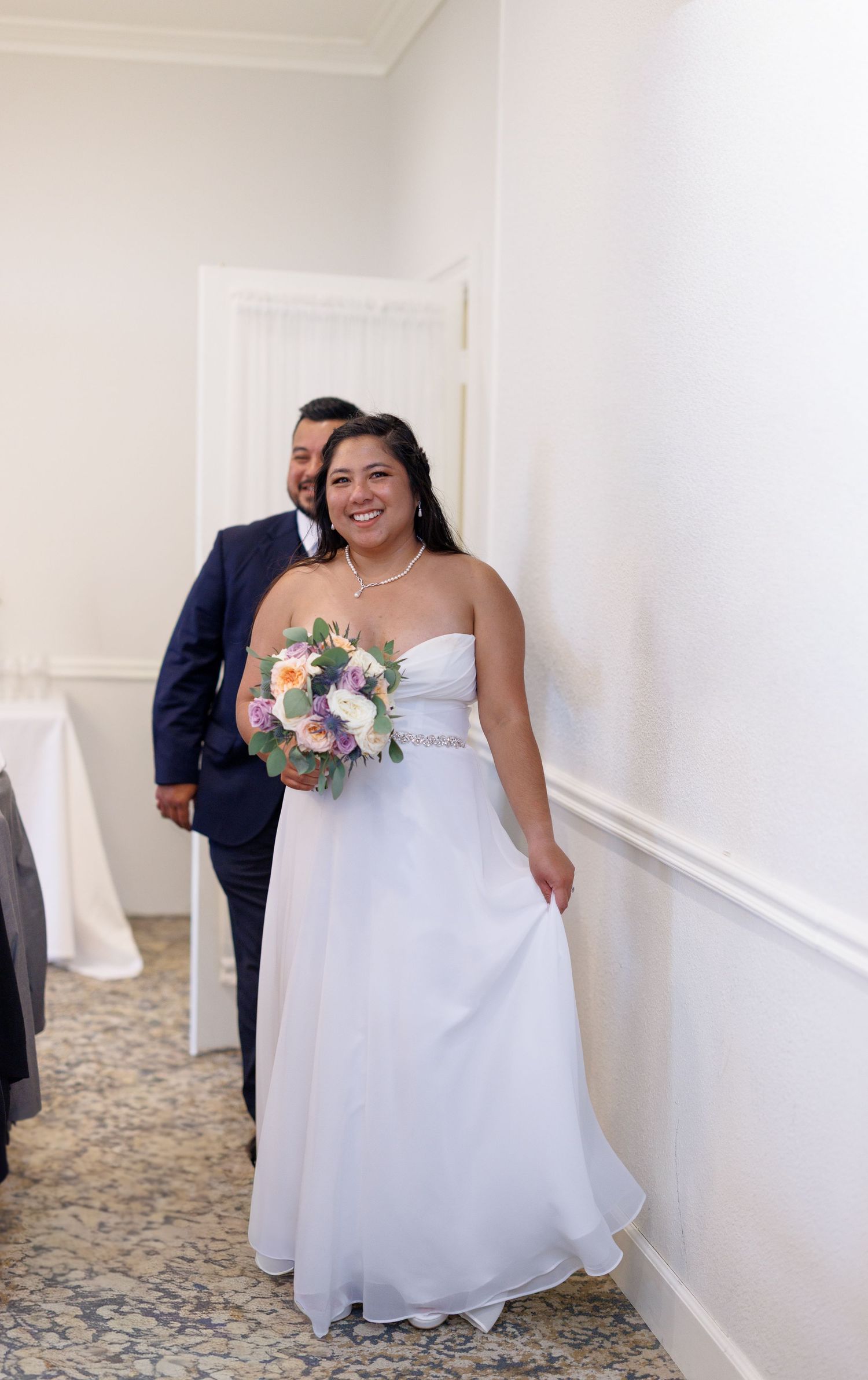 Smiling bride in white strapless gown holding colorful bouquet walks with groom in navy suit at wedding.