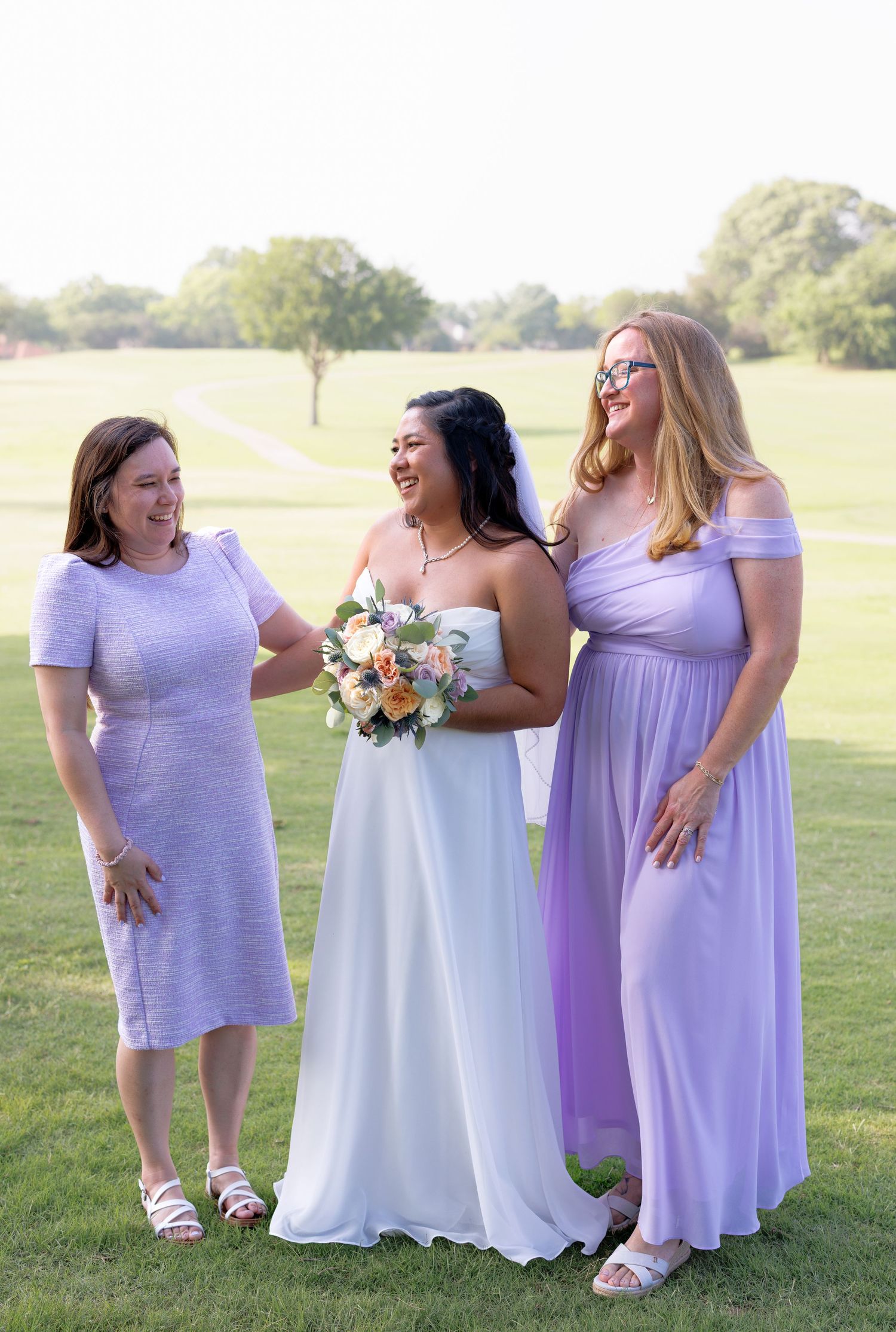 Bride in white gown holding bouquet stands with two women in lavender dresses on a green golf course.