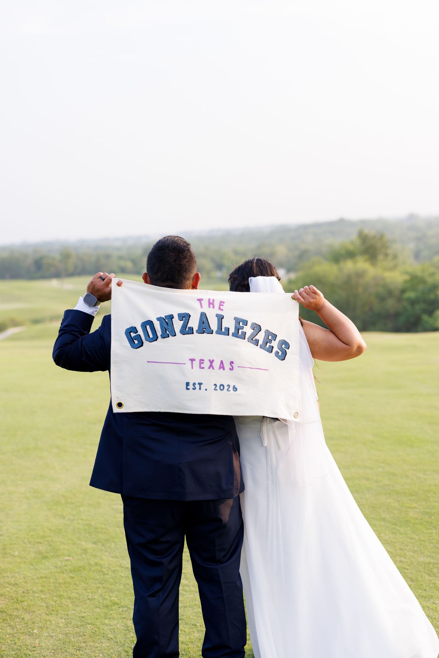 Newlywed couple holding a custom flag reading The Gonzalezes Texas Est. 2026 on a green golf course.