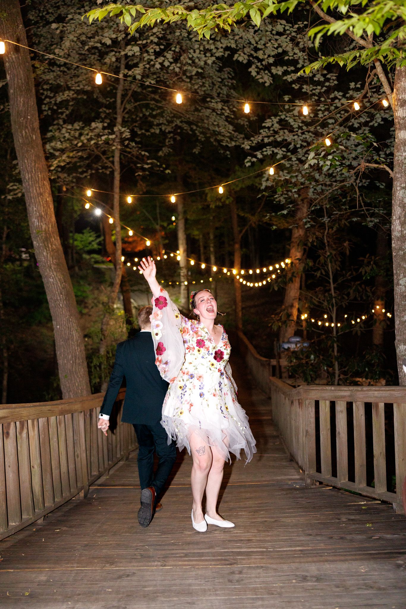 Couple dancing joyfully on a wooden bridge at night, surrounded by string lights and lush trees.