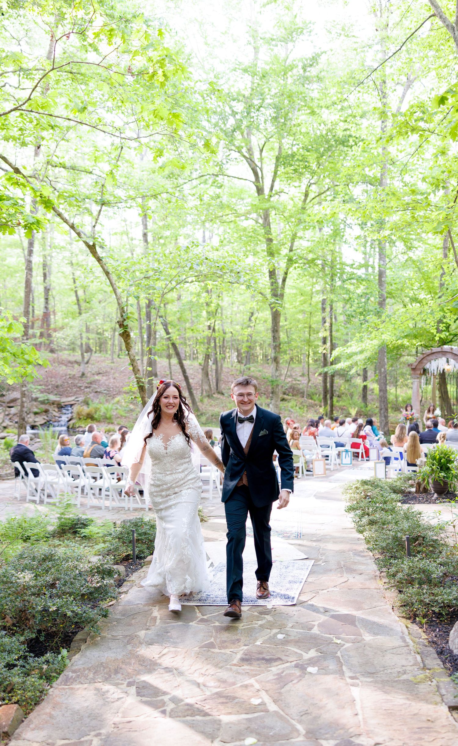 Bride and groom walk hand-in-hand down stone aisle at outdoor woodland wedding ceremony with guests seated among trees.