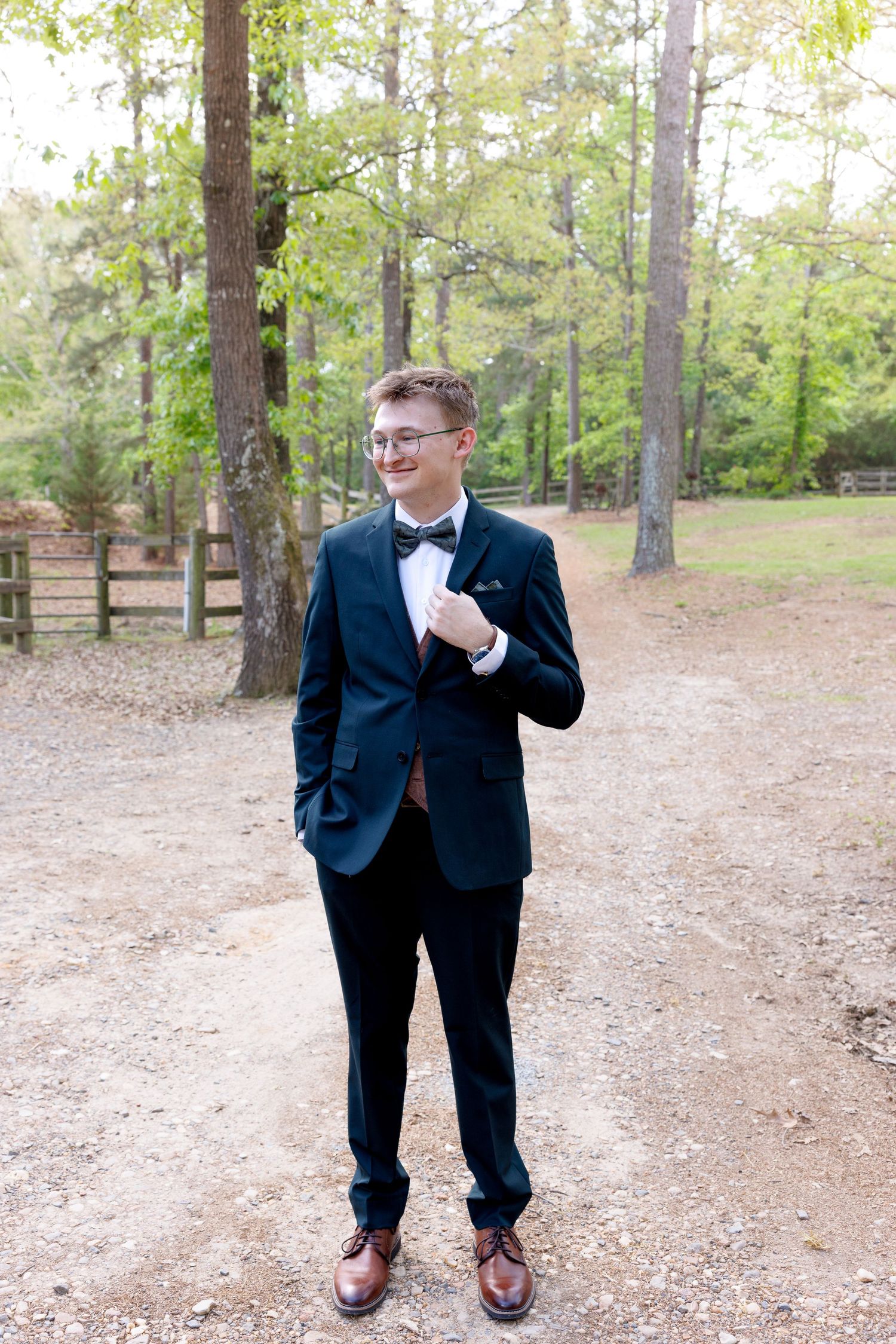 Young man in navy tuxedo with bow tie and glasses posing on gravel path surrounded by tall pine trees.