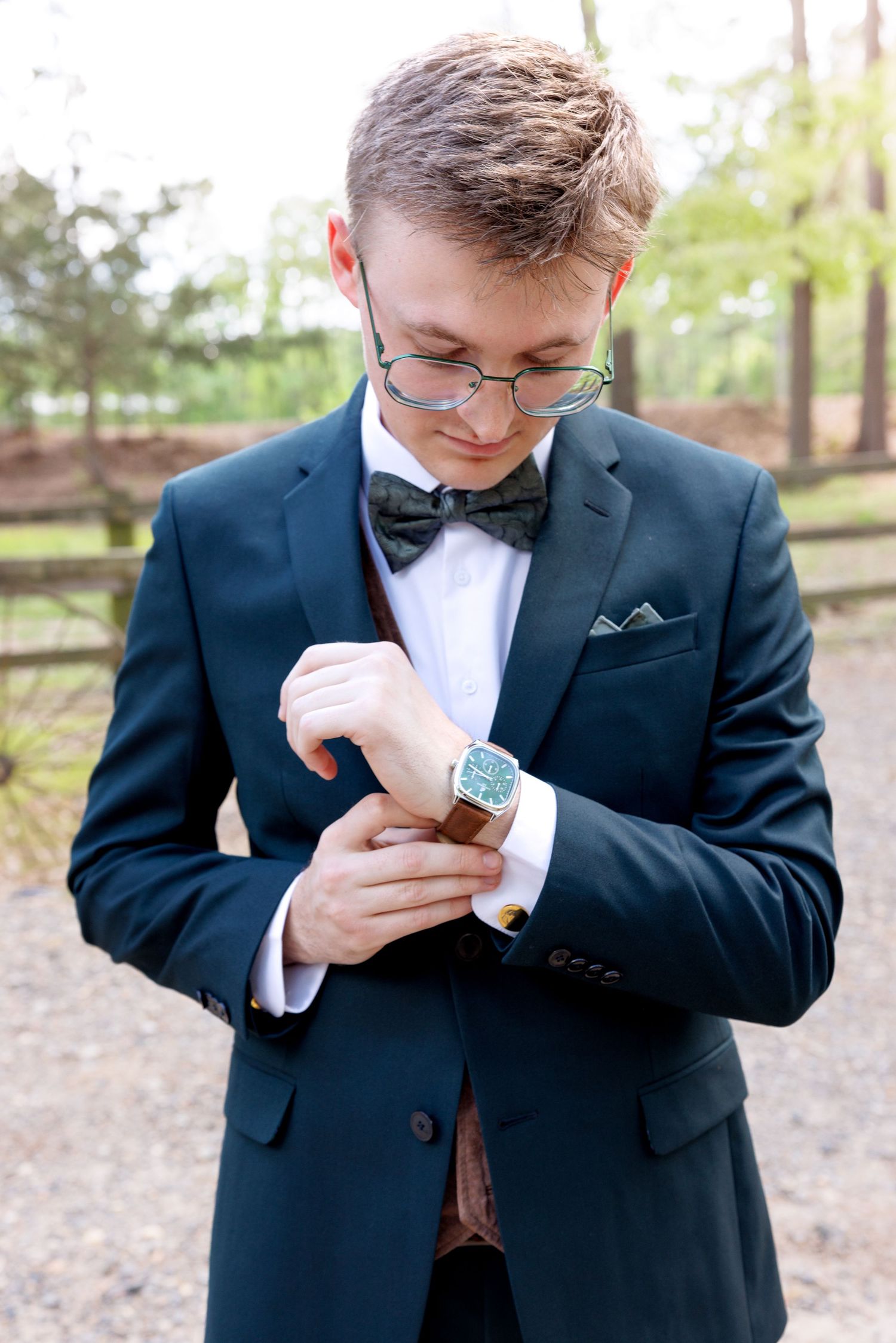 Young man in navy suit with bow tie and glasses adjusting his watch cufflinks outdoors.