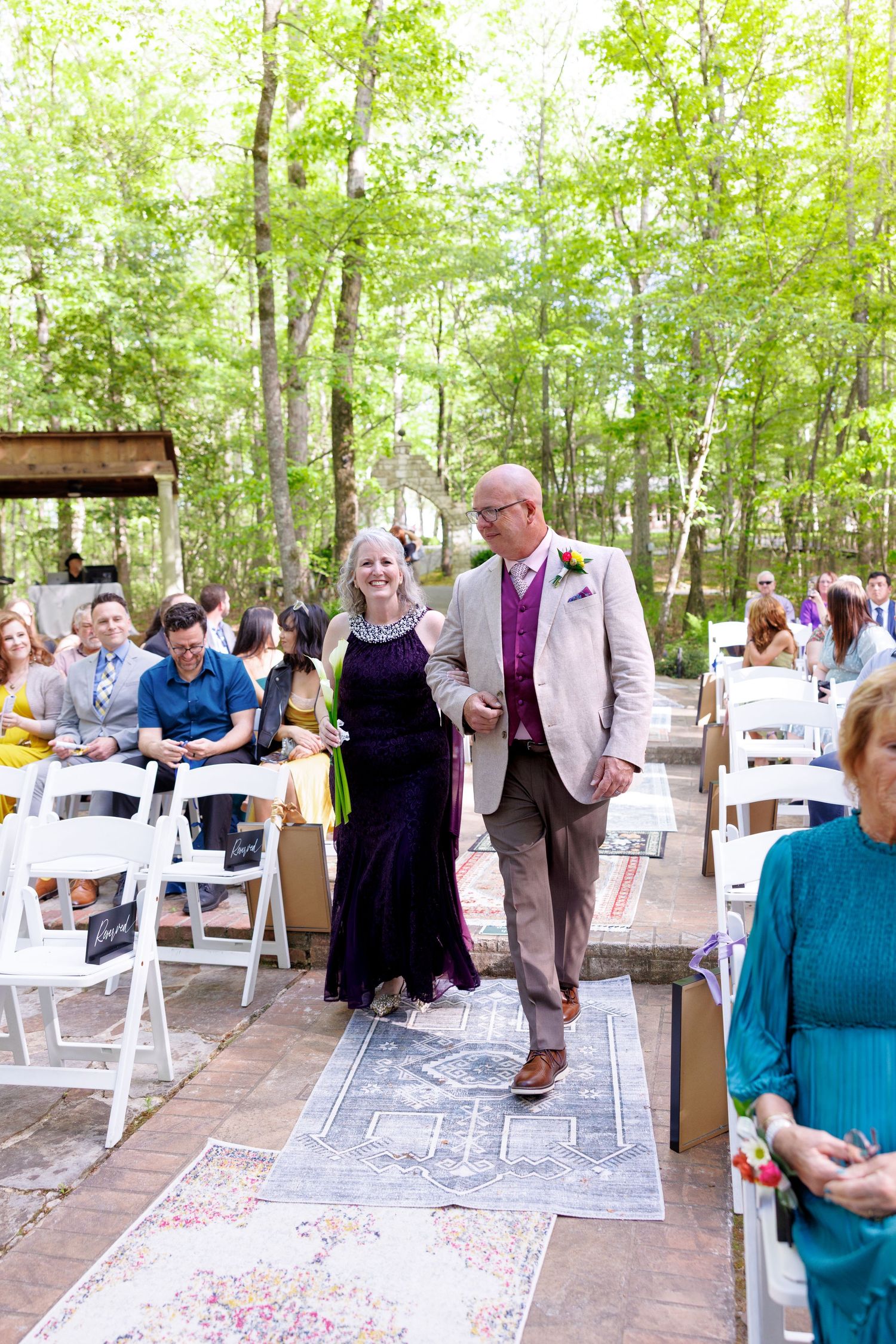 Elegant outdoor wedding processional with guests seated in white chairs amid lush green trees.