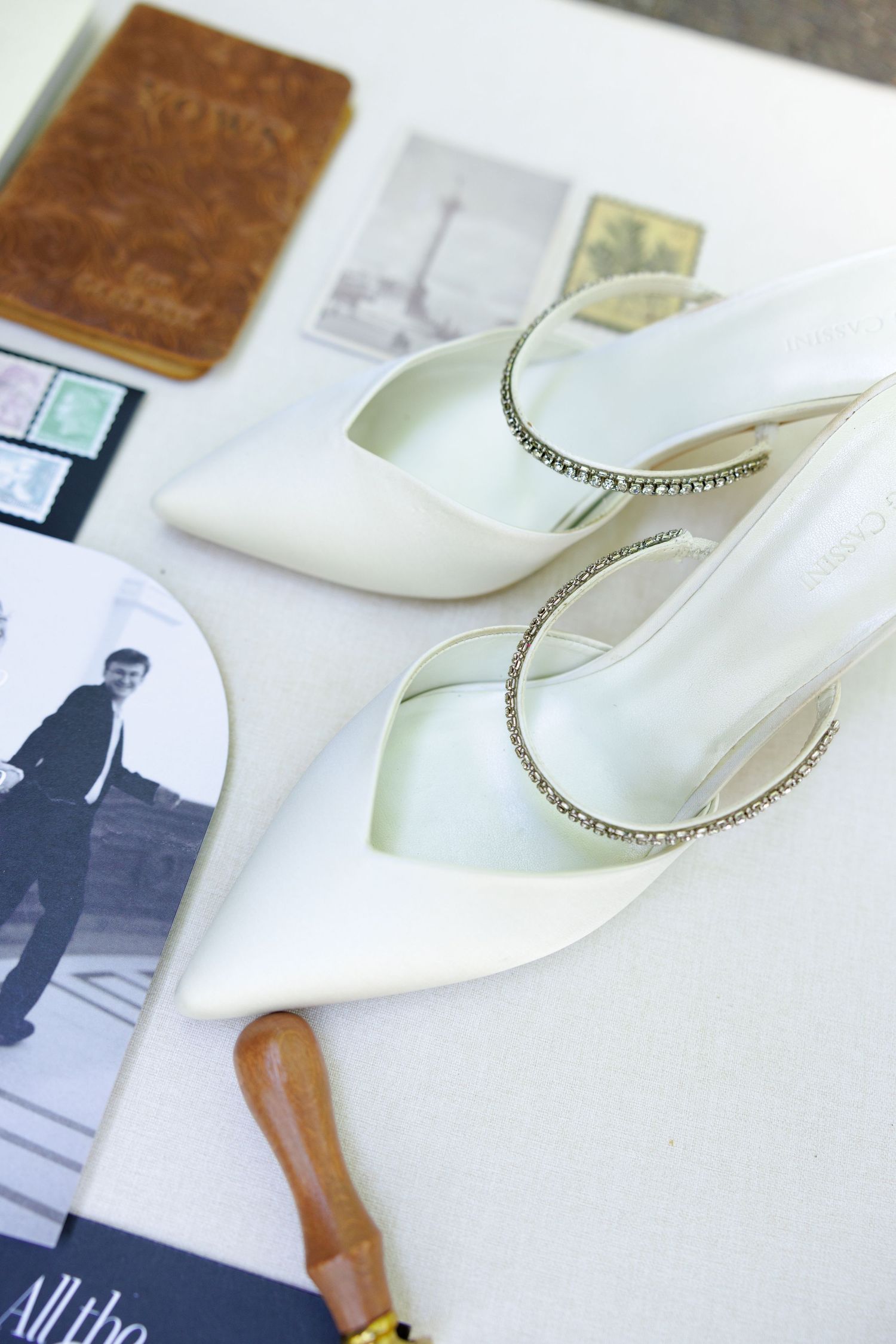 White satin bridal heels with crystal ankle straps displayed on a table with vintage postcards and a leather journal.