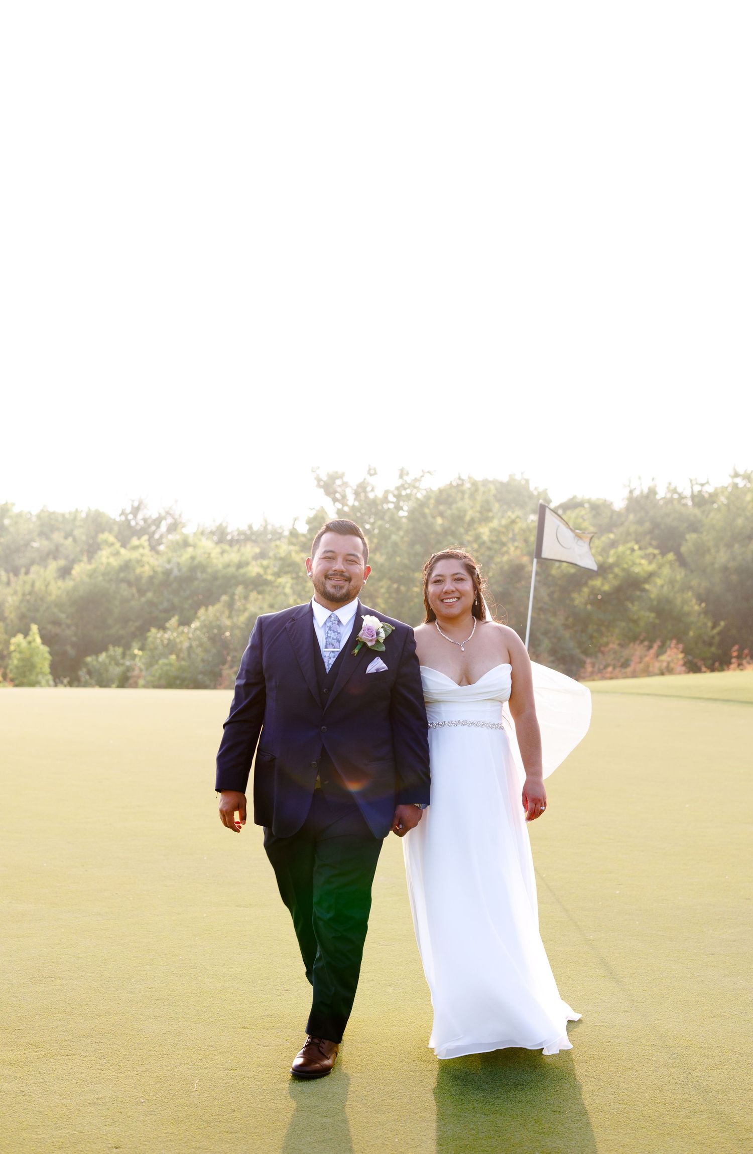 Bride and groom walking hand in hand on a golf course green, smiling, with a white flag and trees in the background.