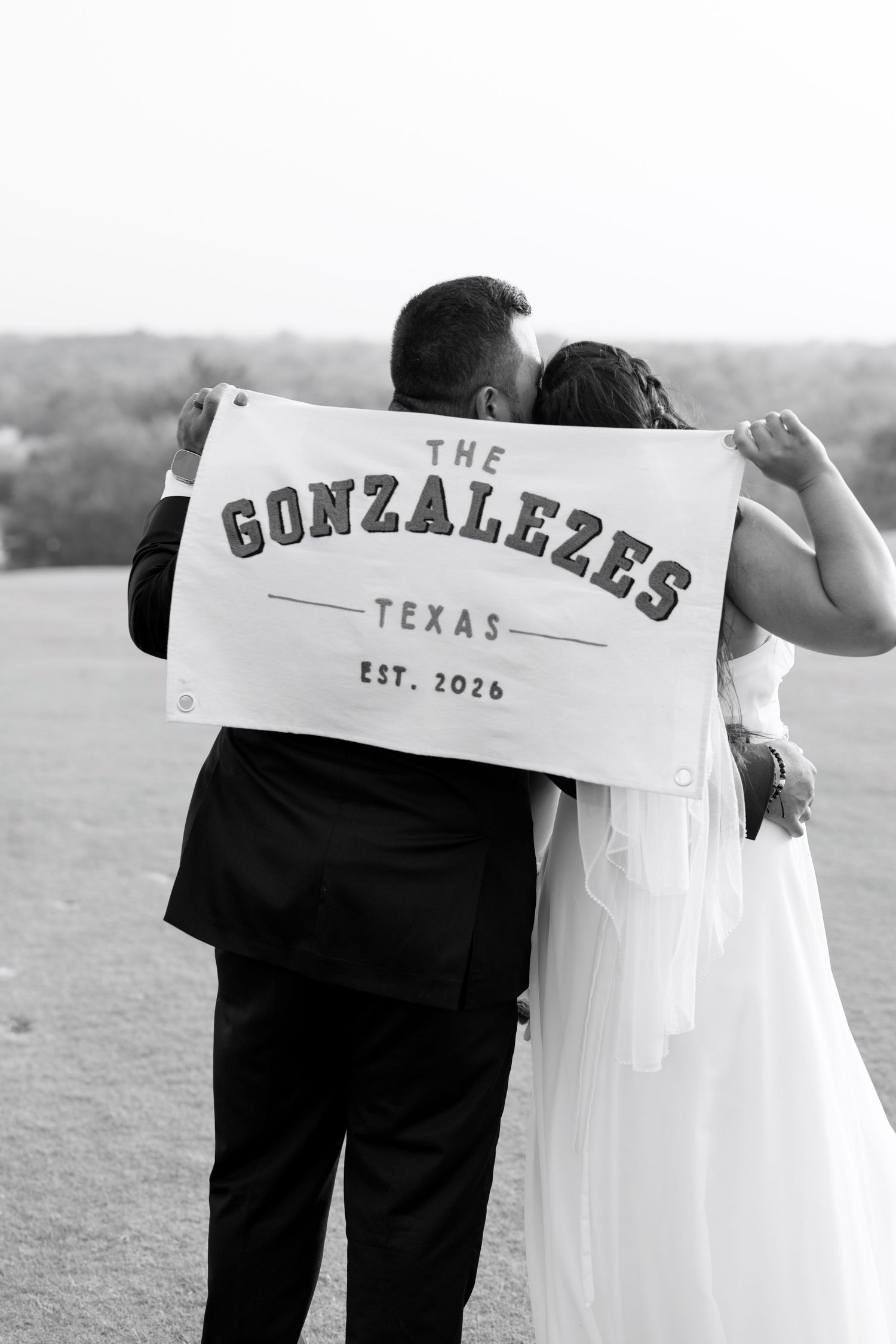 Bride and groom kissing behind a The Gonzalezes Texas Est. 2026 banner in a black and white wedding photo.