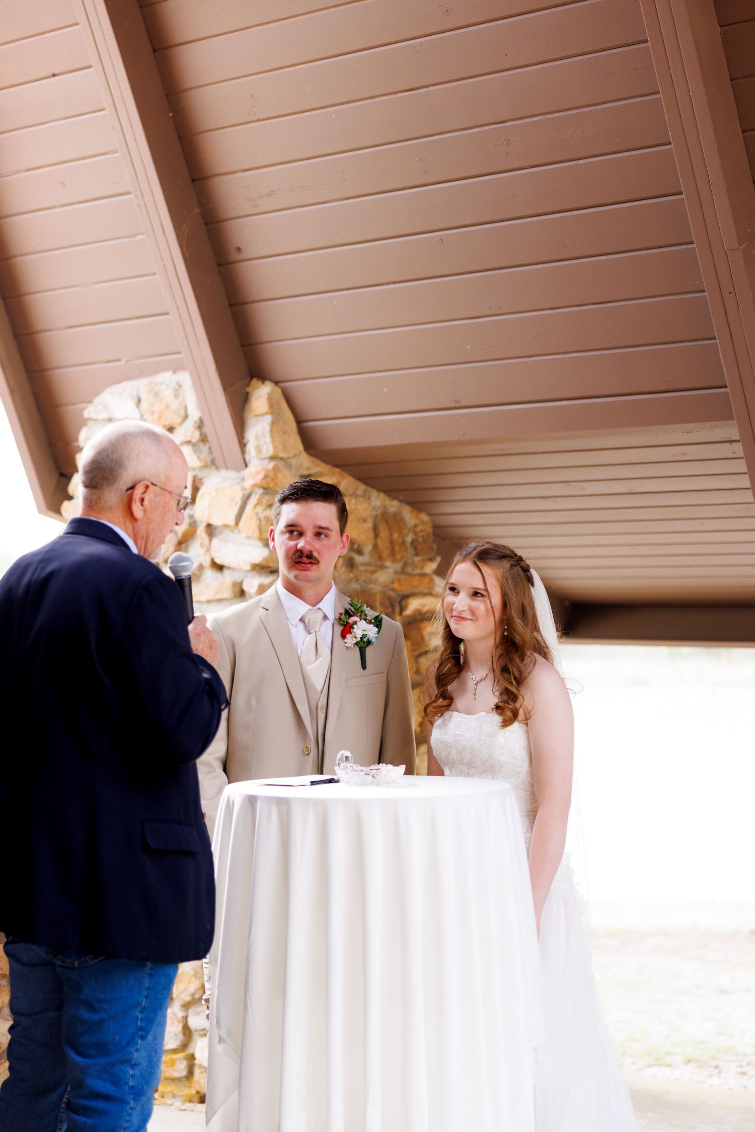 Bride and groom exchange vows with officiant at outdoor wedding ceremony under wooden pavilion.
