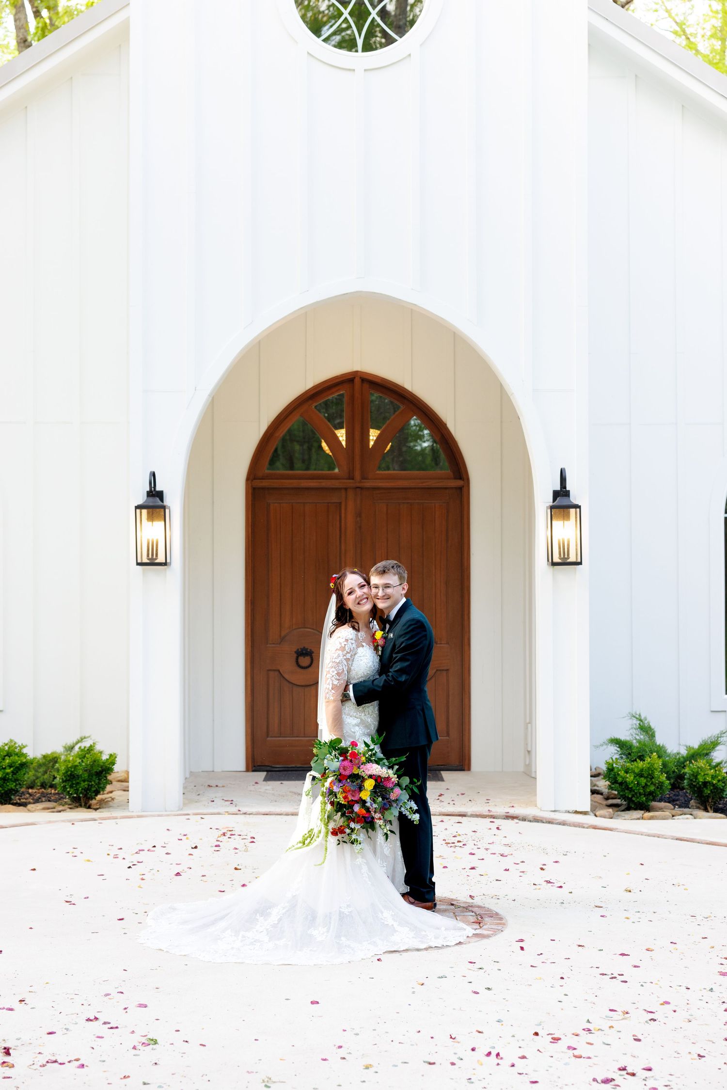Bride and groom pose together in front of a white chapel with wooden doors and rose petals on the ground.
