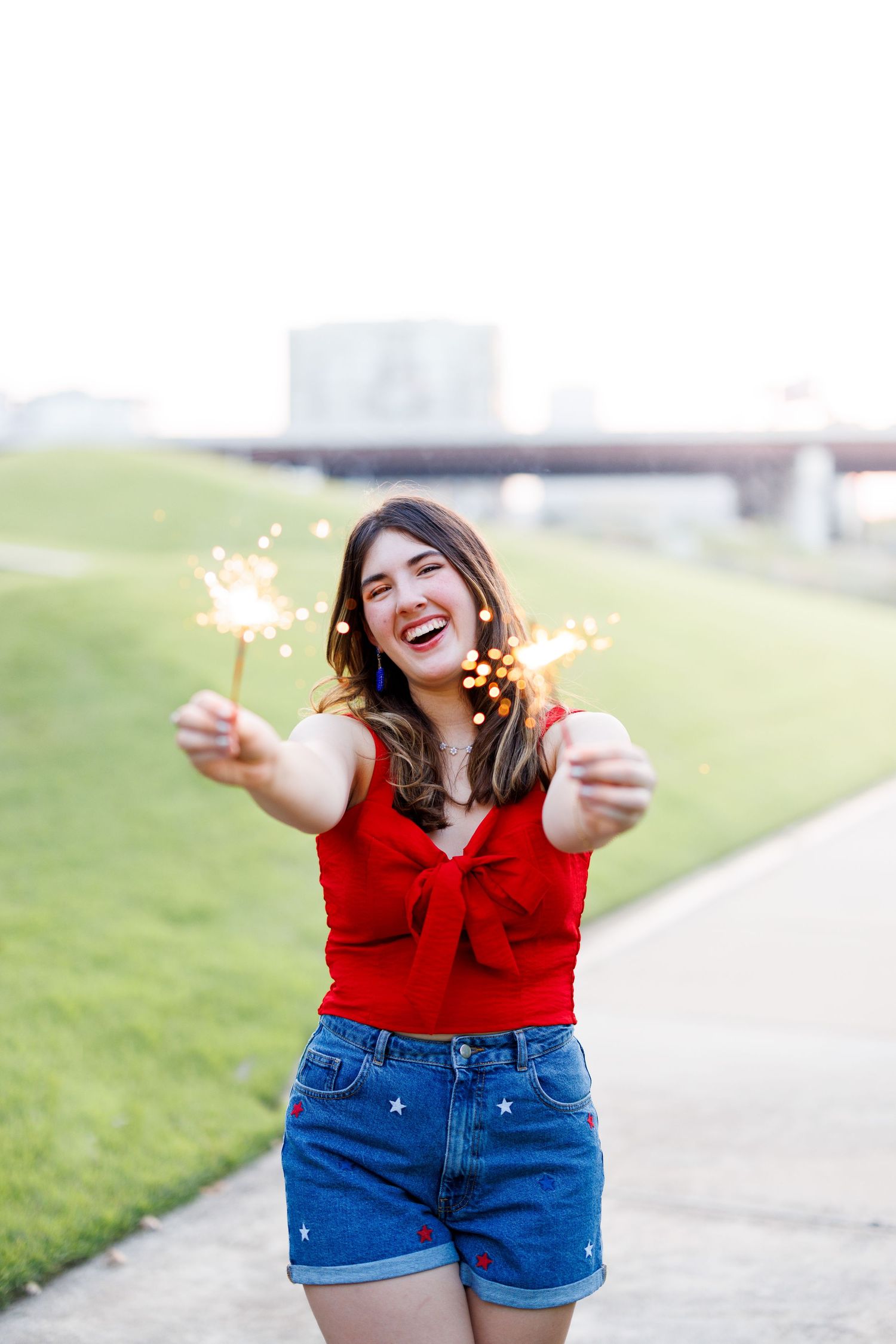 Person in casual summer outfit laughing while holding sparklers on a paved path.