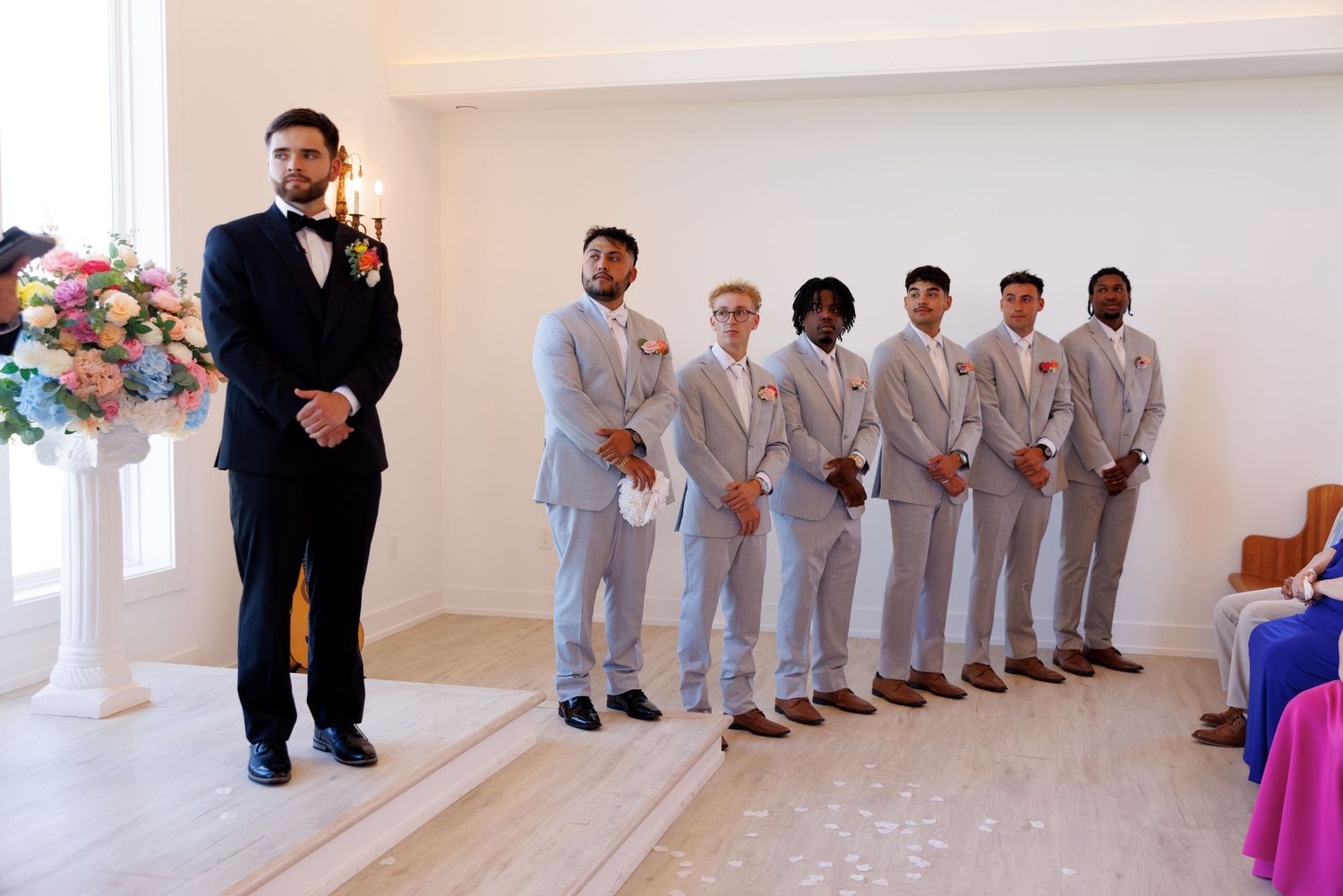 Wedding party members lined up in formal attire against white chapel walls.