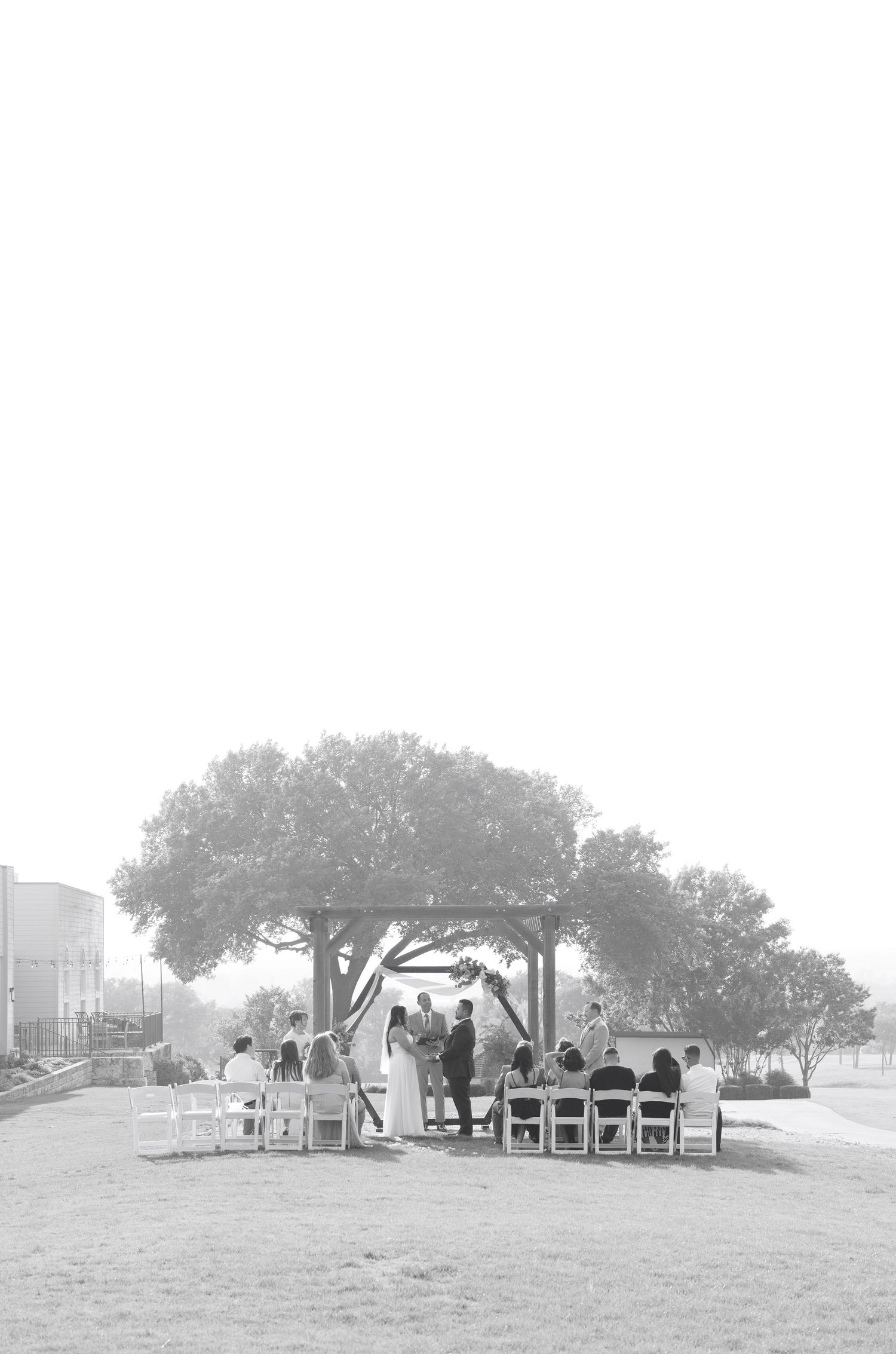 Black and white outdoor wedding ceremony under a pergola surrounded by trees with guests seated in white chairs.