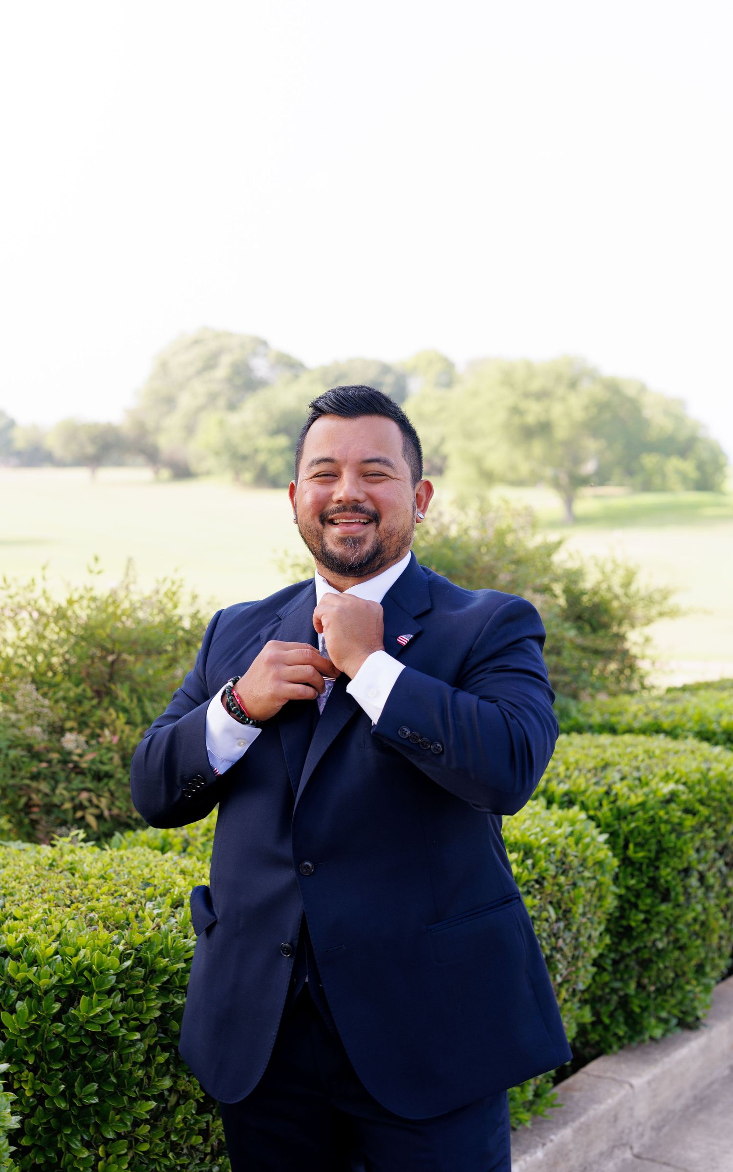 Smiling man in navy suit adjusting tie outdoors with green hedges and trees behind him.