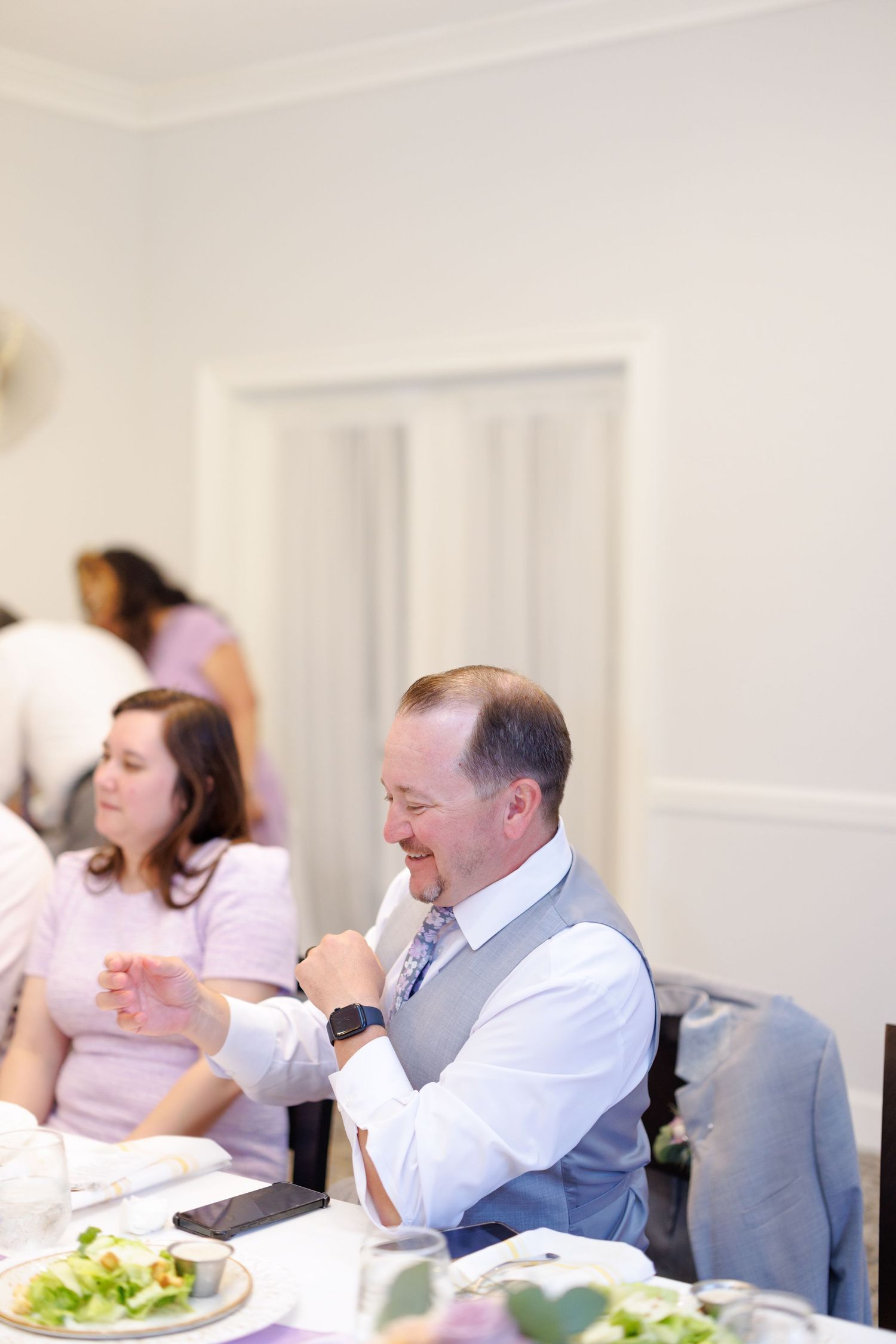 Man in vest and tie smiling at a wedding reception dinner table with guests in background.