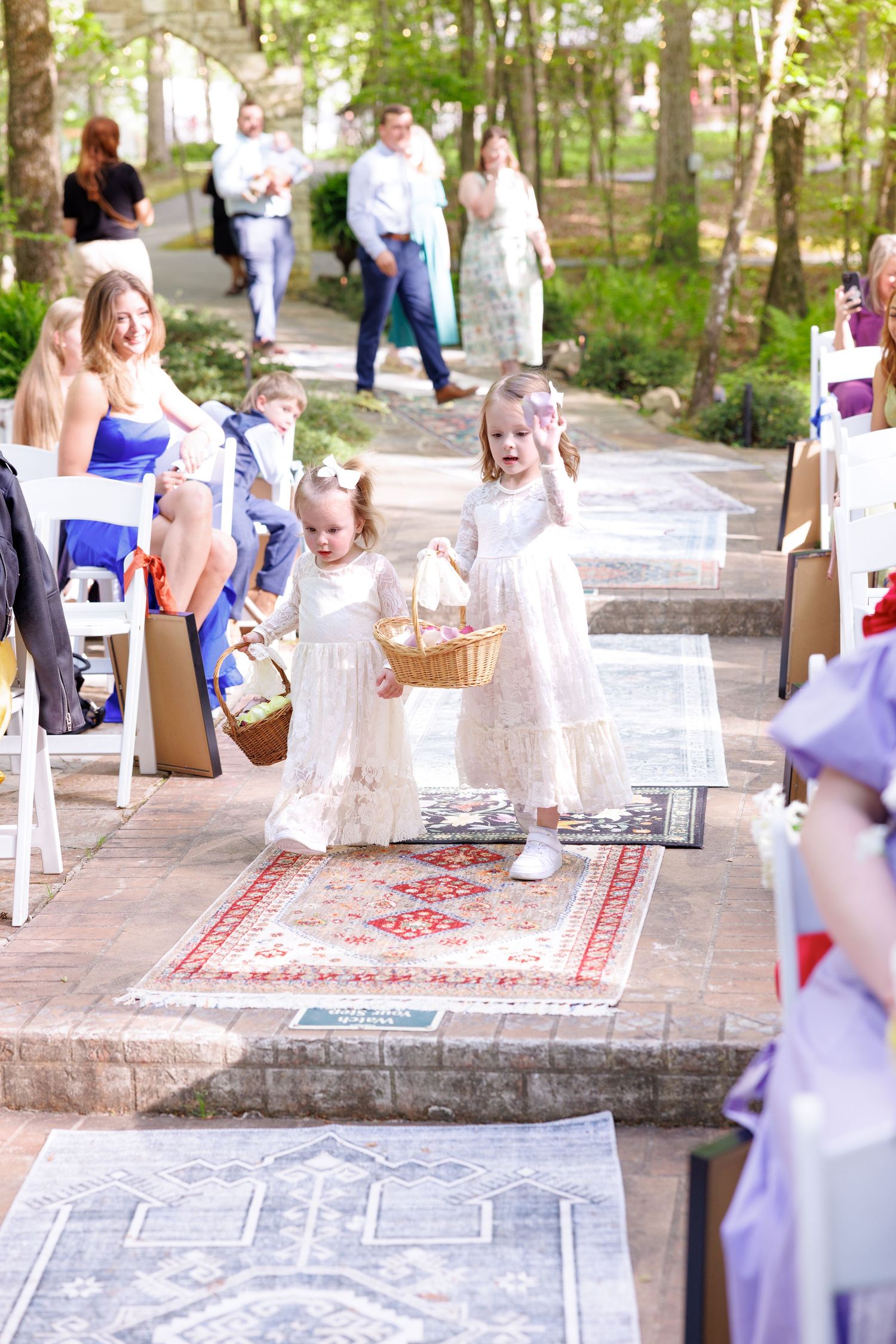Two flower girls in white dresses walk down an outdoor wedding aisle carrying a basket.