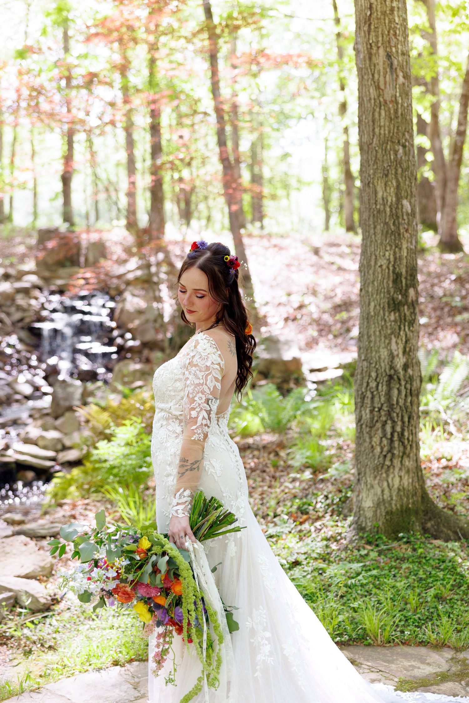Bride in long-sleeve lace wedding gown holding colorful bouquet, standing in lush forest near waterfall.