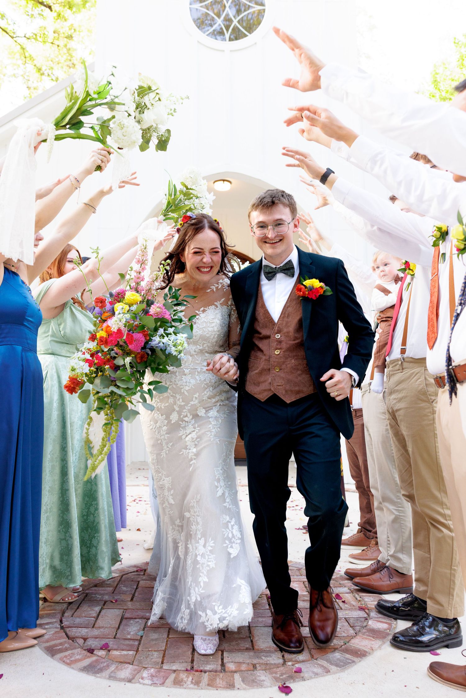 Bride and groom exit church through tunnel of raised hands, bride holding colorful bouquet, guests celebrating outdoors.