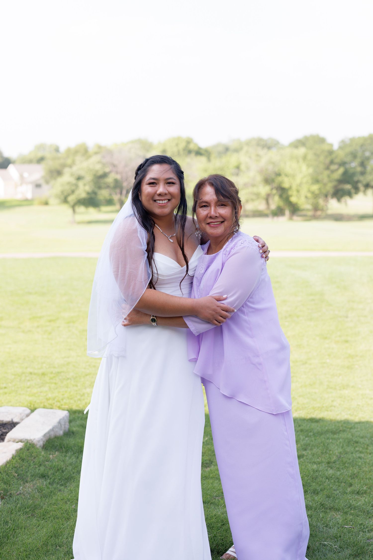 Bride in white gown and veil hugging woman in lavender dress outdoors on green lawn at wedding celebration.