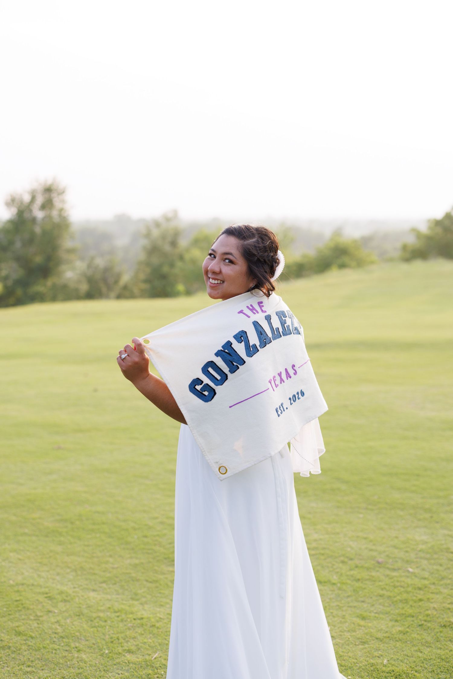 Smiling bride in white dress holding a custom Gonzalez Texas family flag on a green golf course.