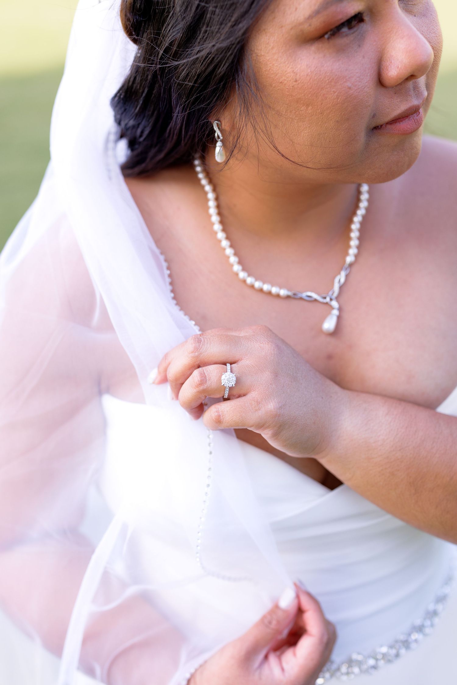 Bride in white gown adjusting veil, wearing pearl necklace and diamond engagement ring on her wedding day.