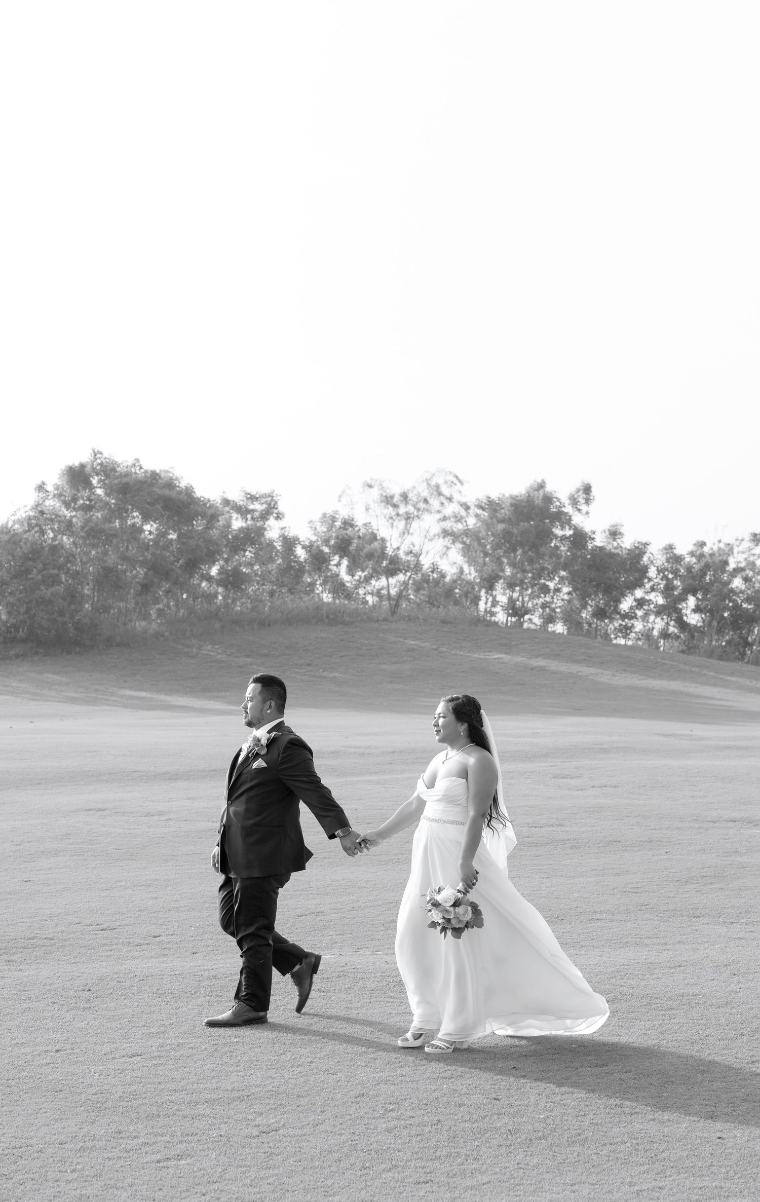 Black and white photo of a bride and groom holding hands walking across an open field on their wedding day.