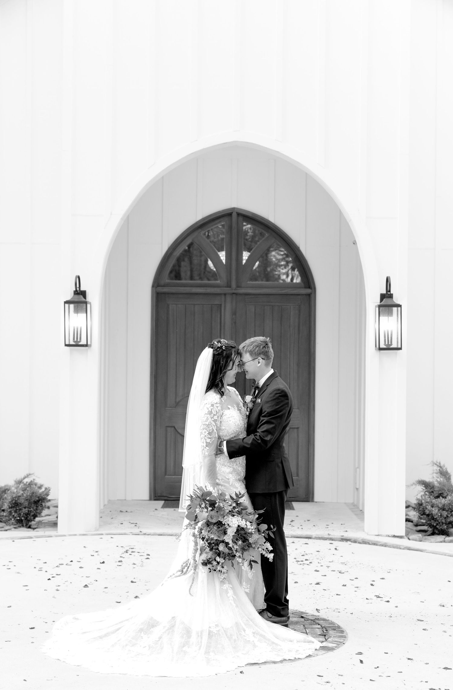 Black and white photo of bride and groom embracing before elegant white chapel with arched wooden doors.