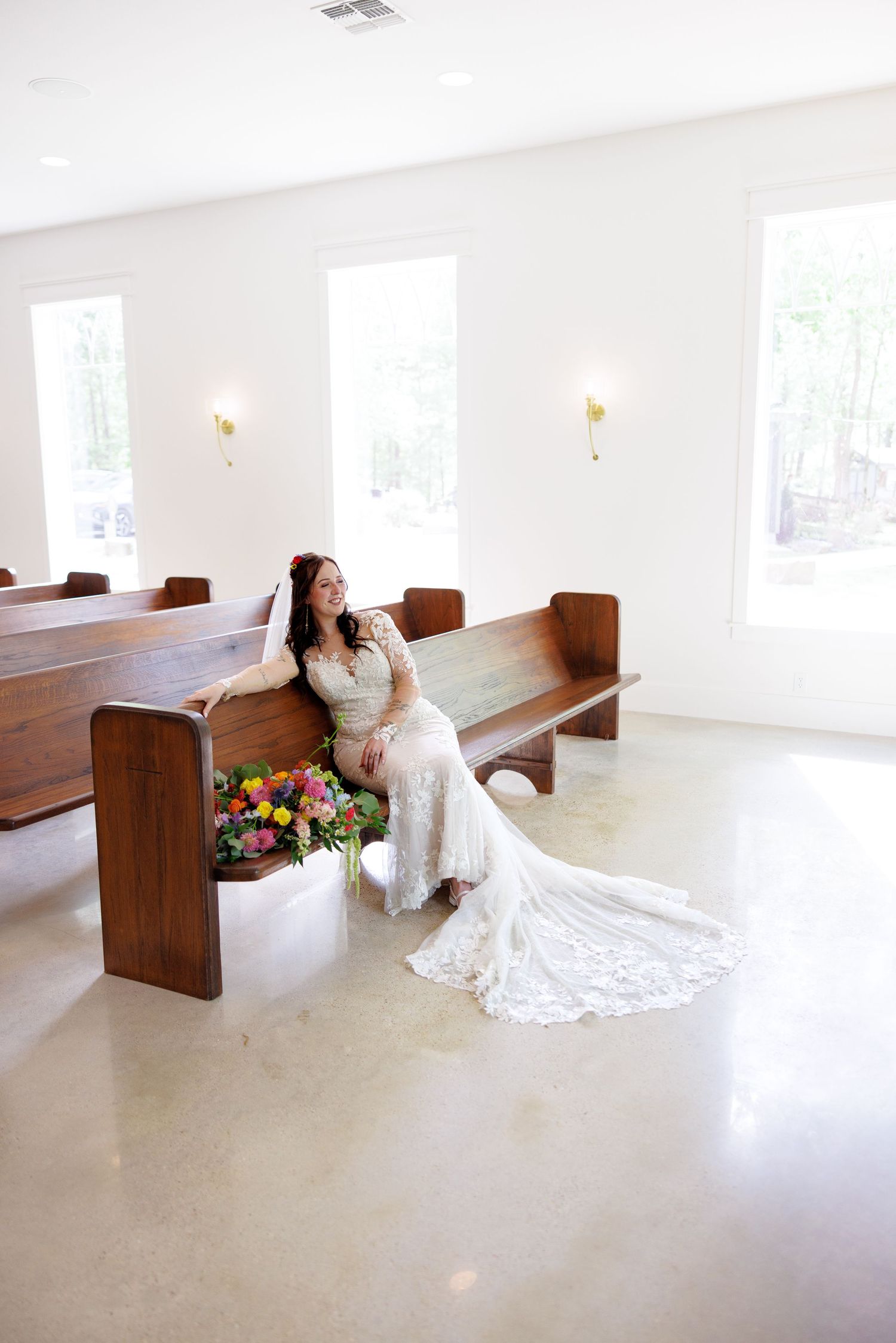 Bride in lace wedding gown sits on wooden church pew with colorful bridal bouquet in bright white chapel.