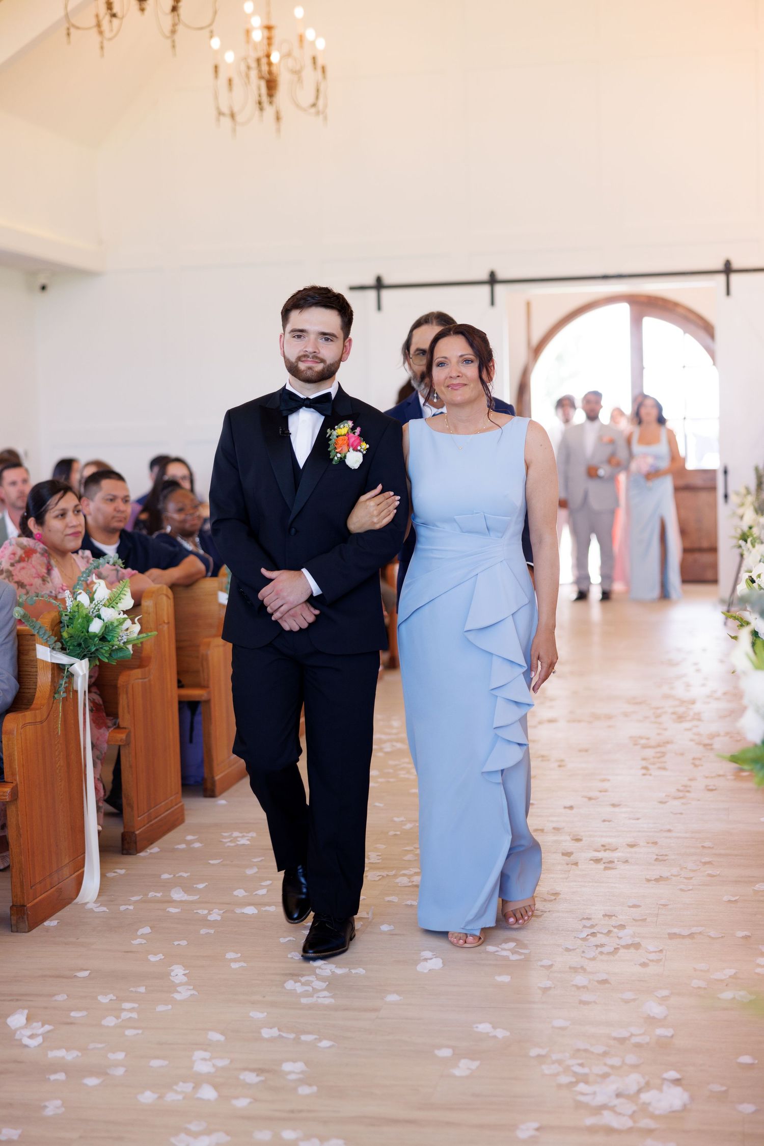 Wedding party walks down church aisle decorated with scattered rose petals and wooden pews.