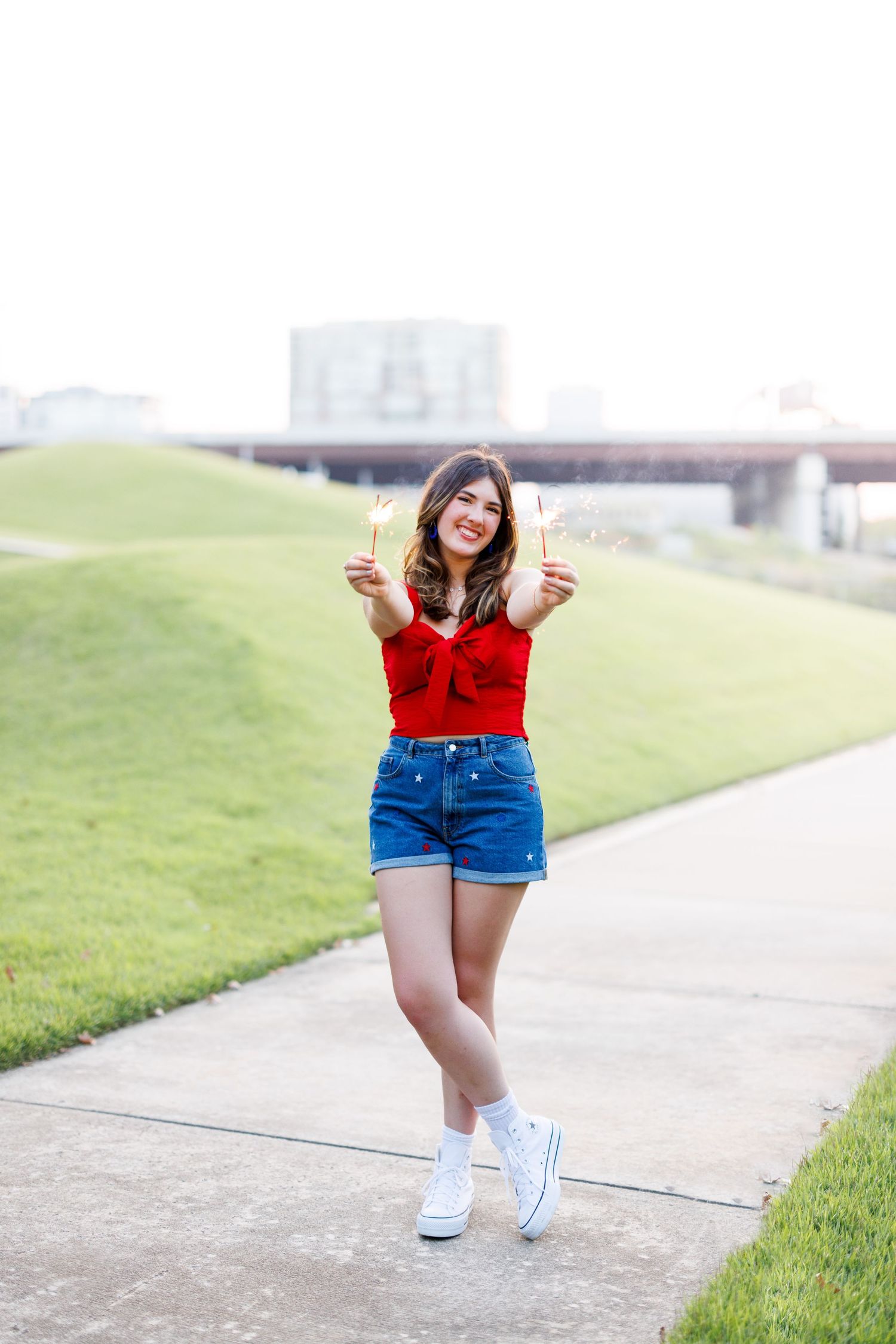 Person wearing red top and blue shorts dancing with sparklers on a grassy hill.