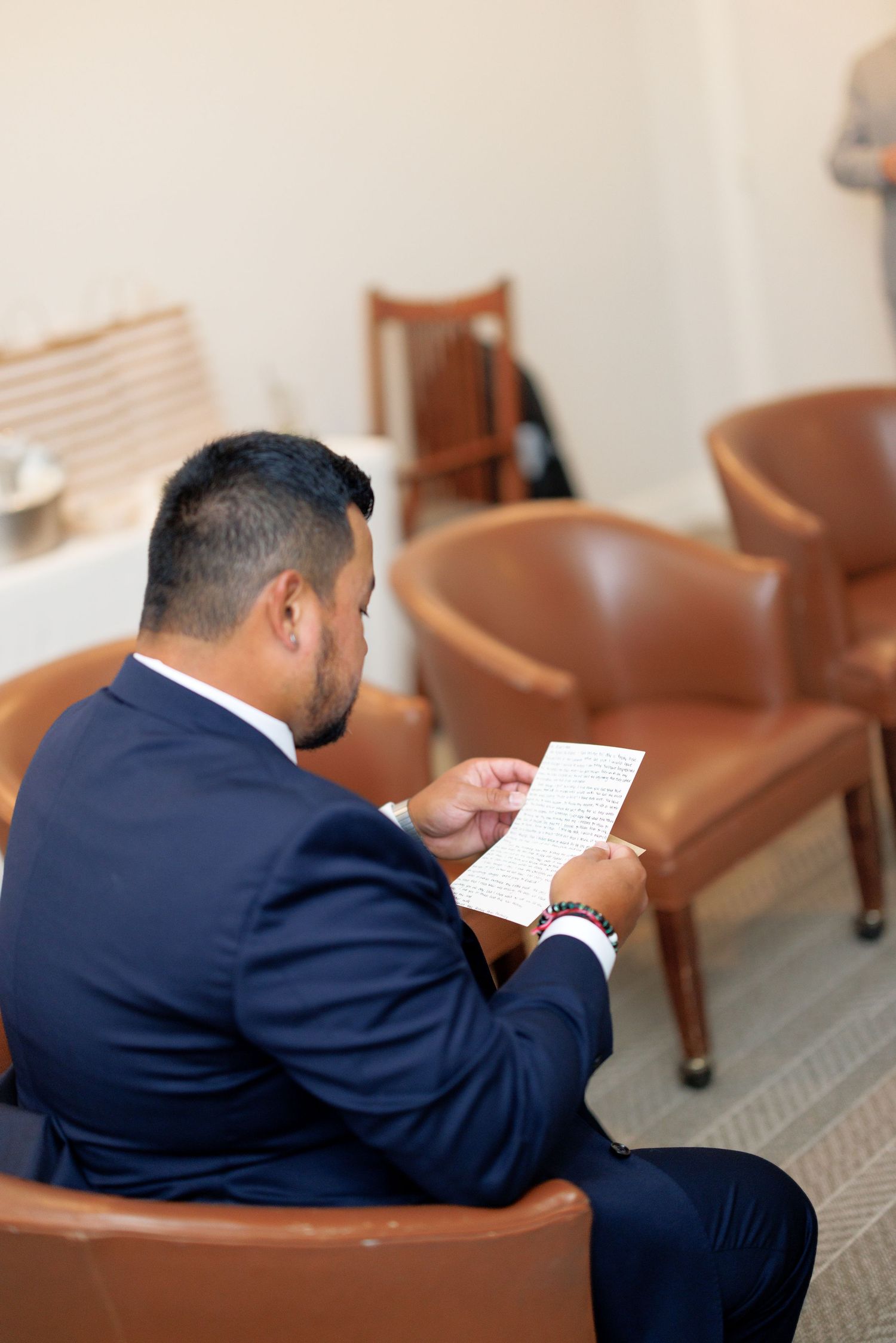 Man in navy suit sitting in brown leather chair reading a handwritten note in a modern waiting room.