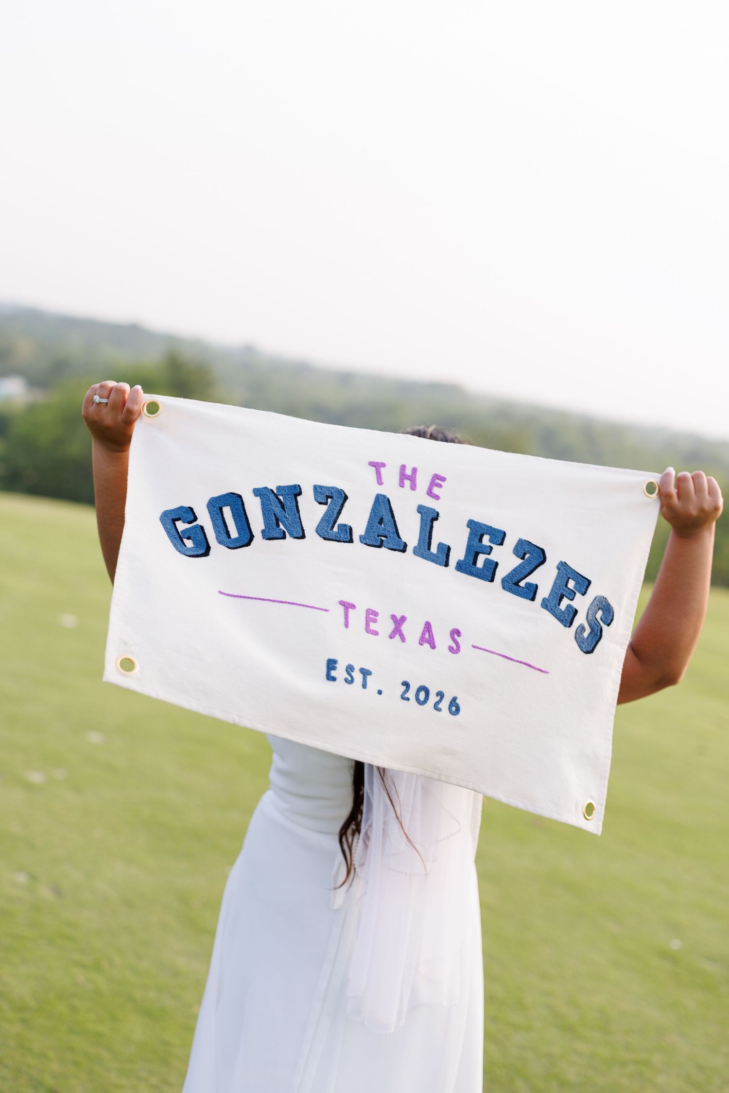 Person holding a custom white banner reading The Gonzalezes Texas Est. 2026 outdoors on a green field.