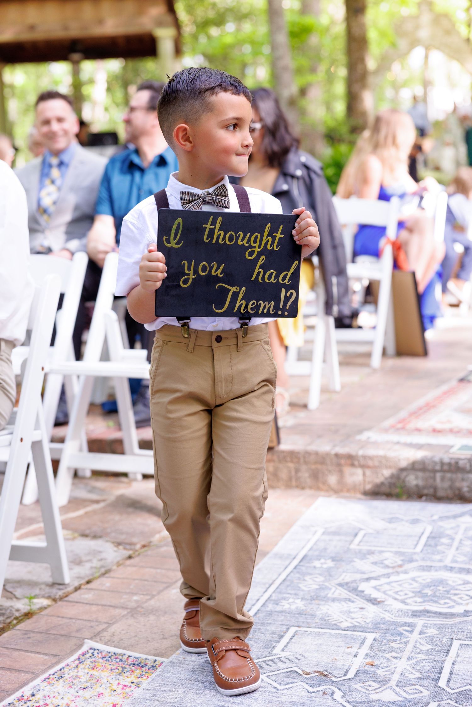 A young ring bearer walks down the wedding aisle holding a chalkboard sign reading I thought you had them at an outdoor ceremony.
