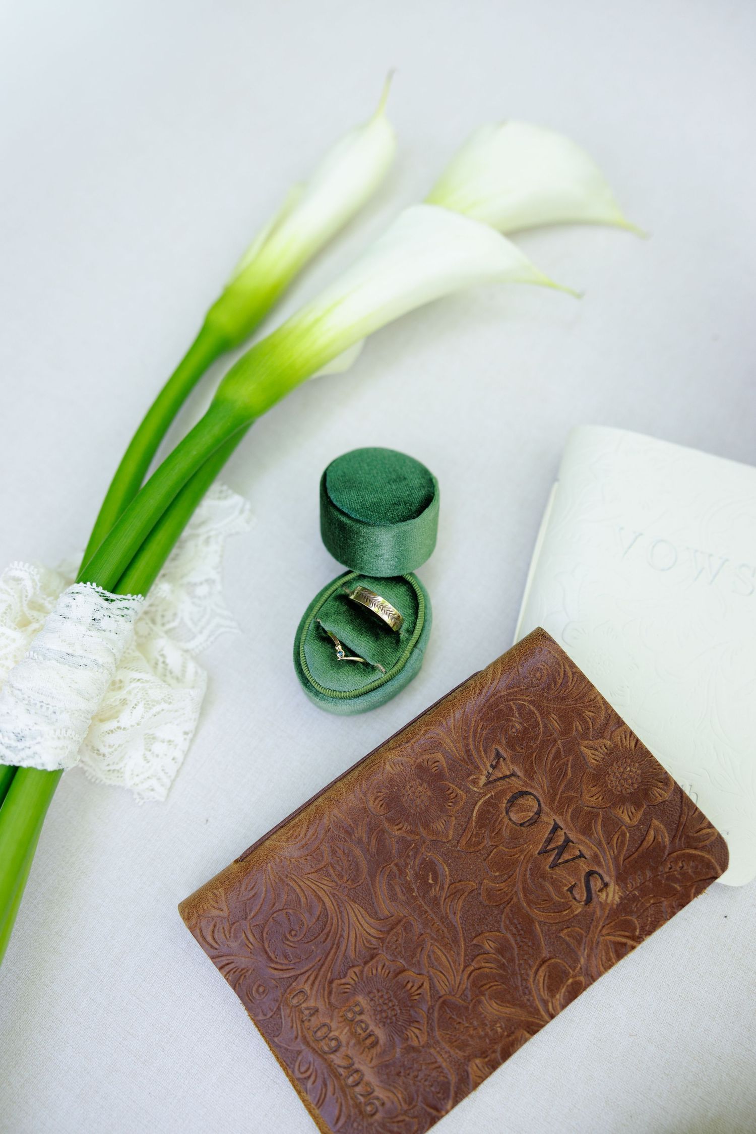 Wedding flat lay with gold rings in green velvet box, calla lilies, lace garter, and leather vows book.