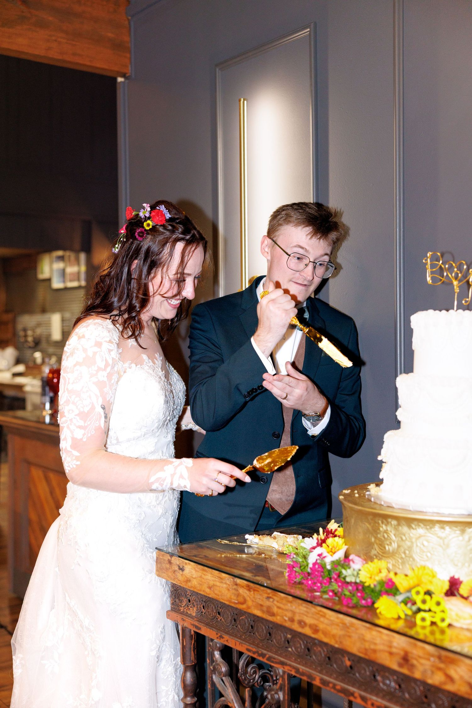 Bride and groom cutting their white wedding cake together, smiling, with floral decorations at an elegant indoor reception.
