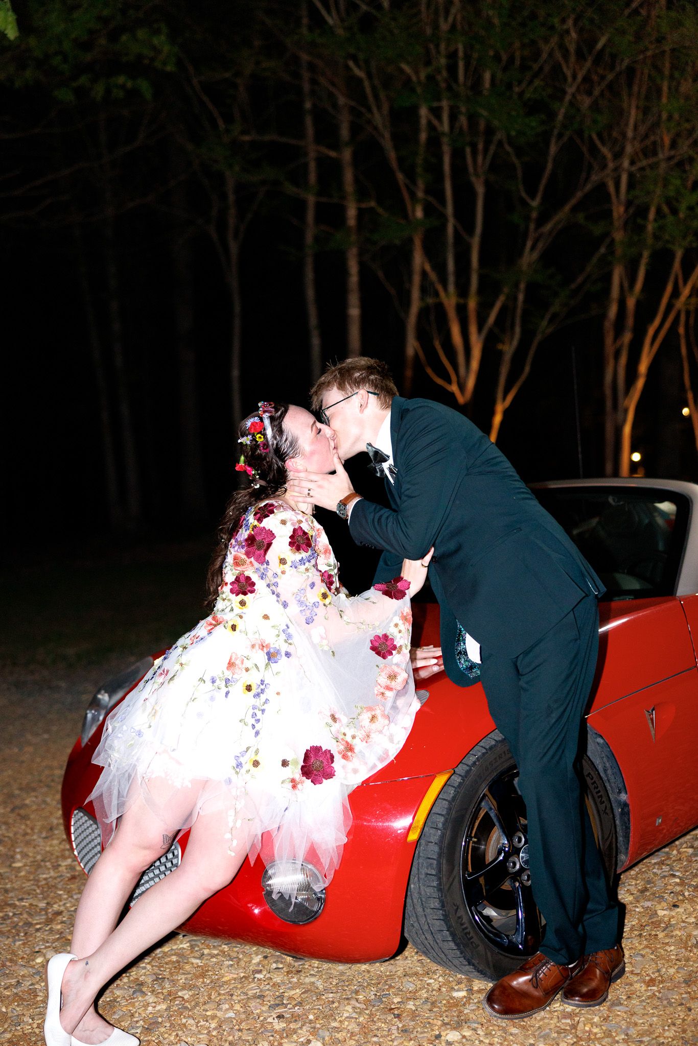 Couple in formal attire kissing on a red sports car at night, woman in floral dress, man in dark suit.