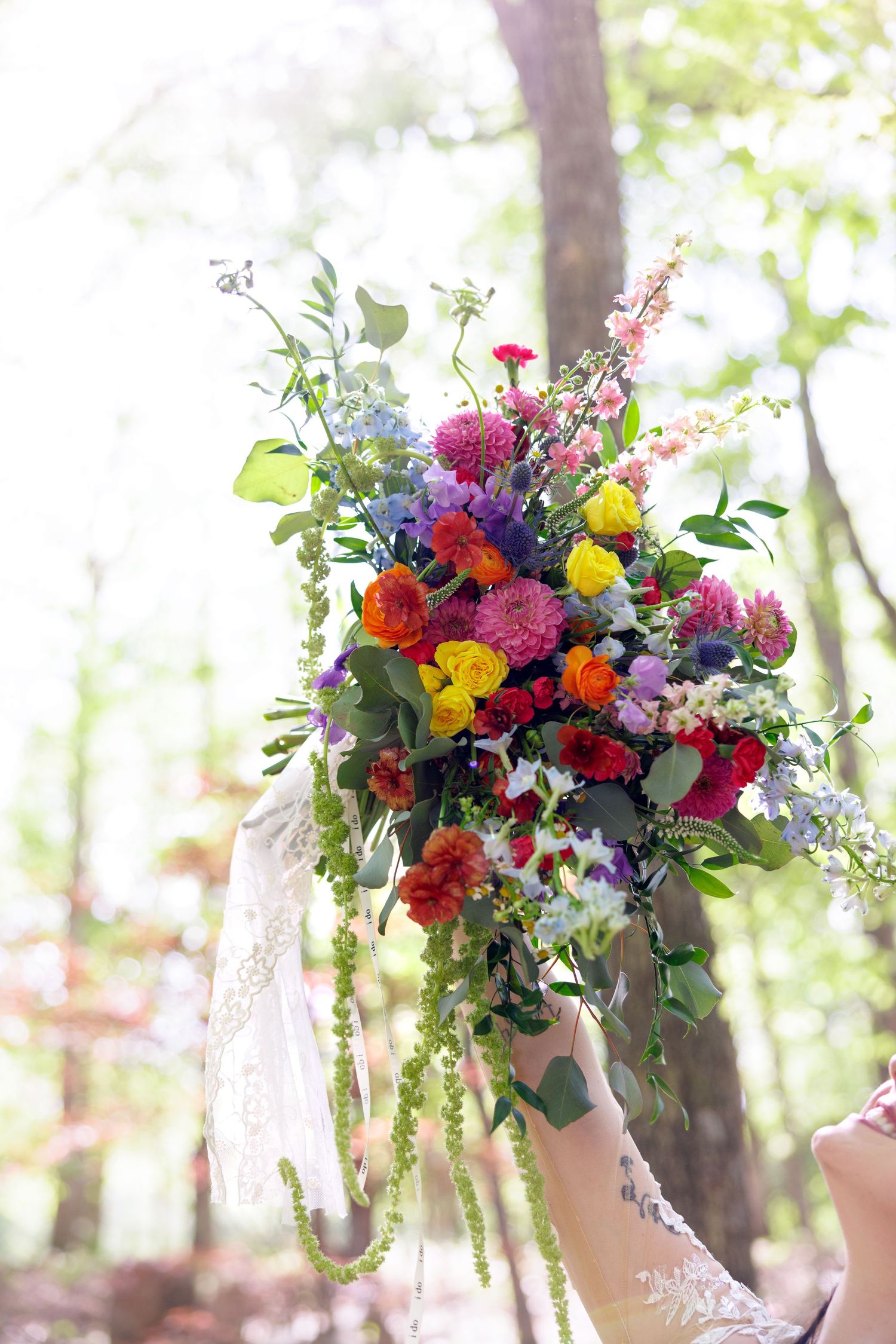 Bride holding a vibrant wildflower wedding bouquet with colorful blooms in a sunlit forest setting.