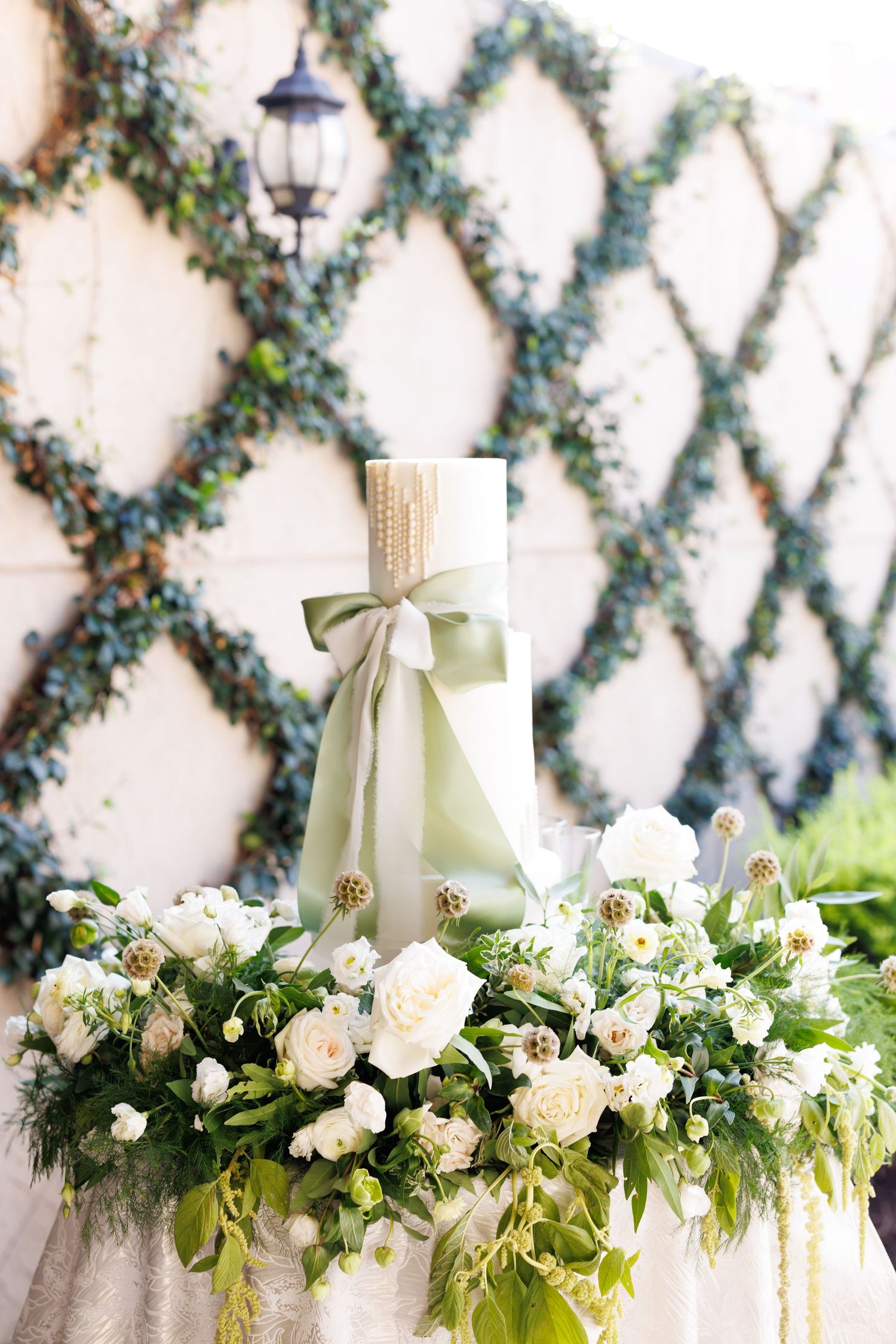 Elegant white floral table decorations with cream ribbons against a latticed garden wall.