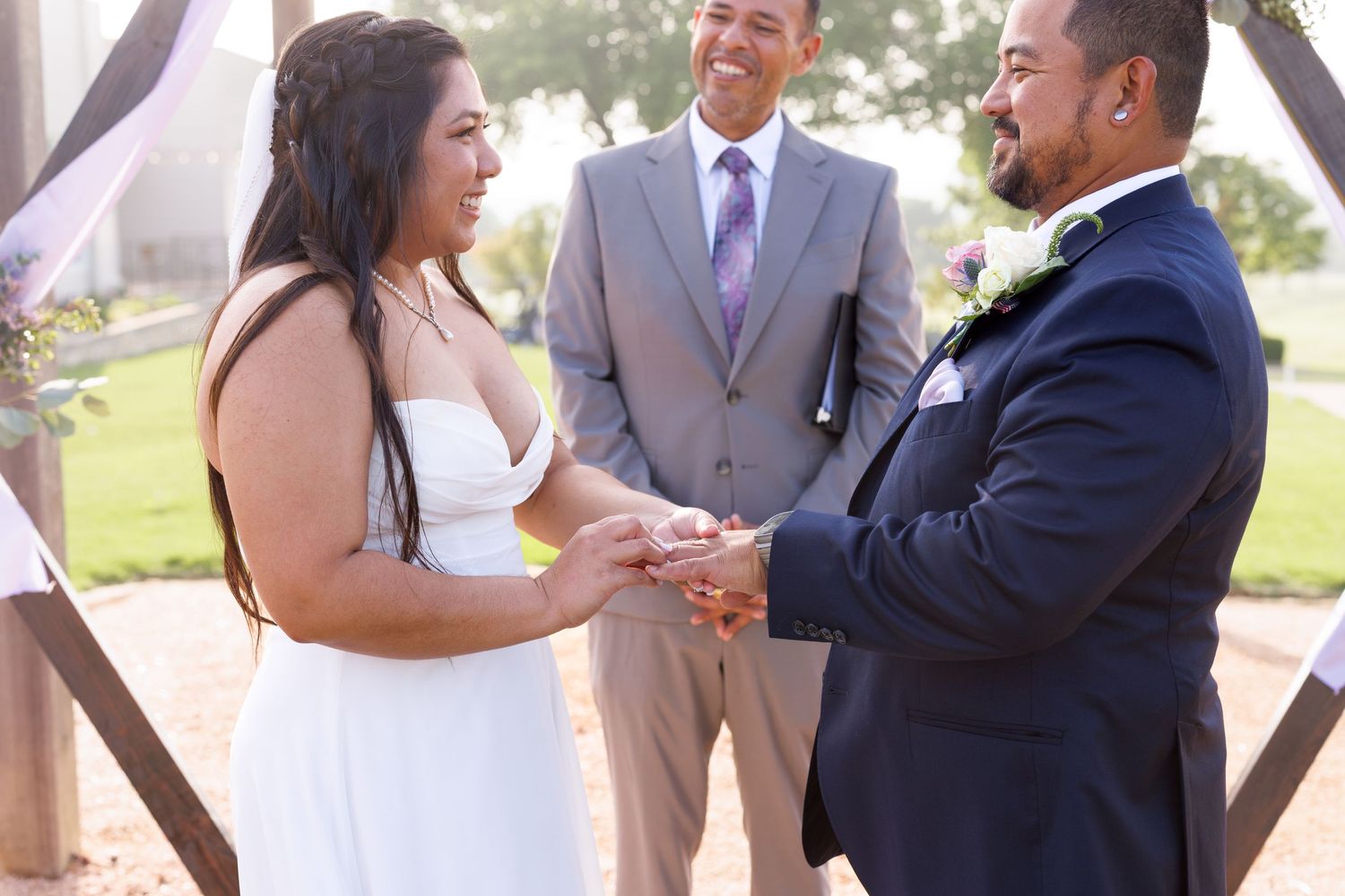 Bride and groom exchanging rings at outdoor wedding ceremony with smiling officiant in background.