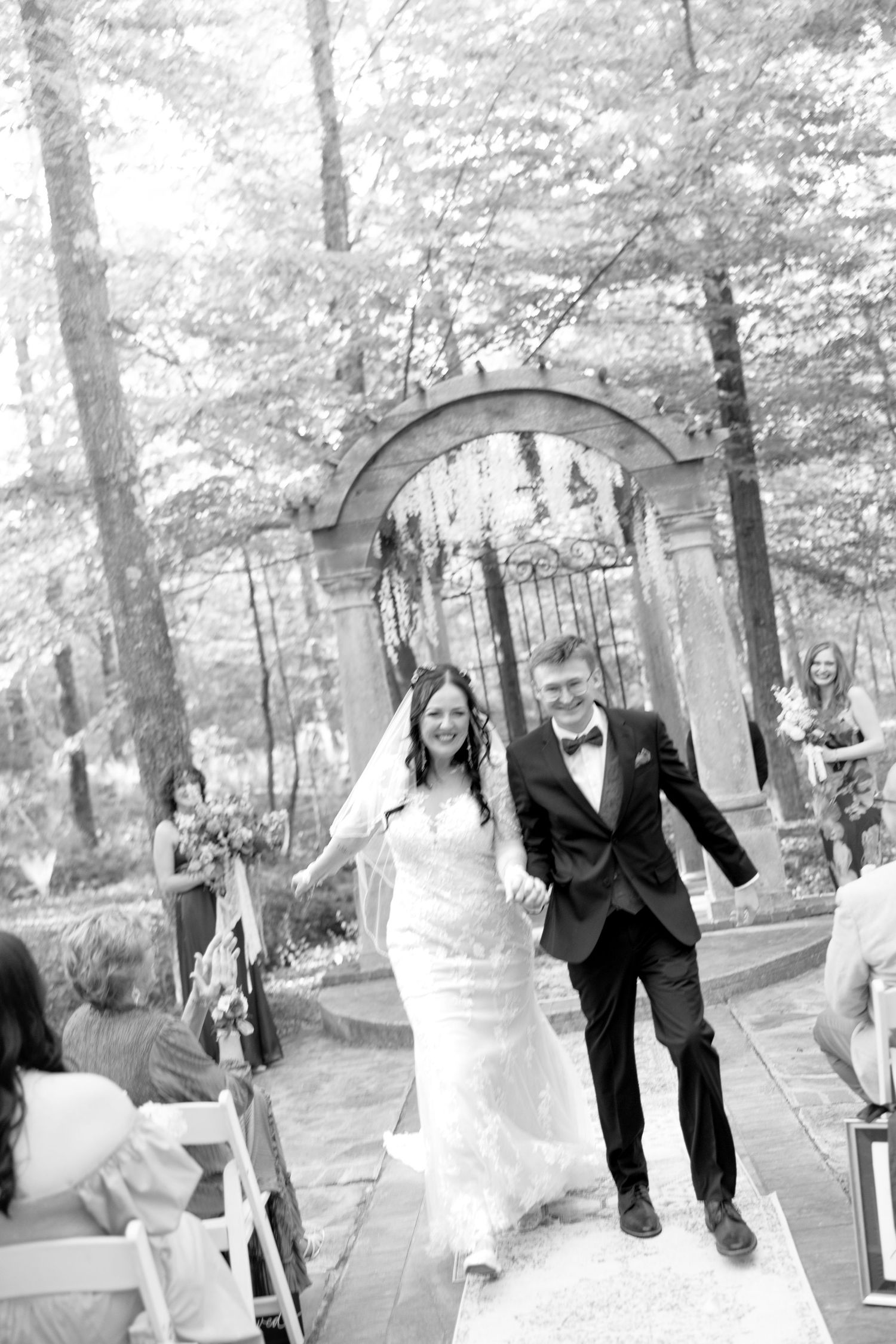 Newlyweds walk joyfully down aisle after outdoor woodland wedding ceremony in black and white photo.
