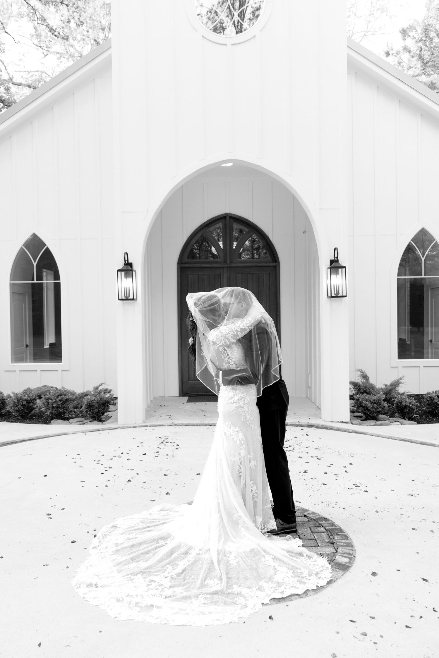 Bride and groom share a romantic kiss under a flowing veil outside a white chapel in black and white.