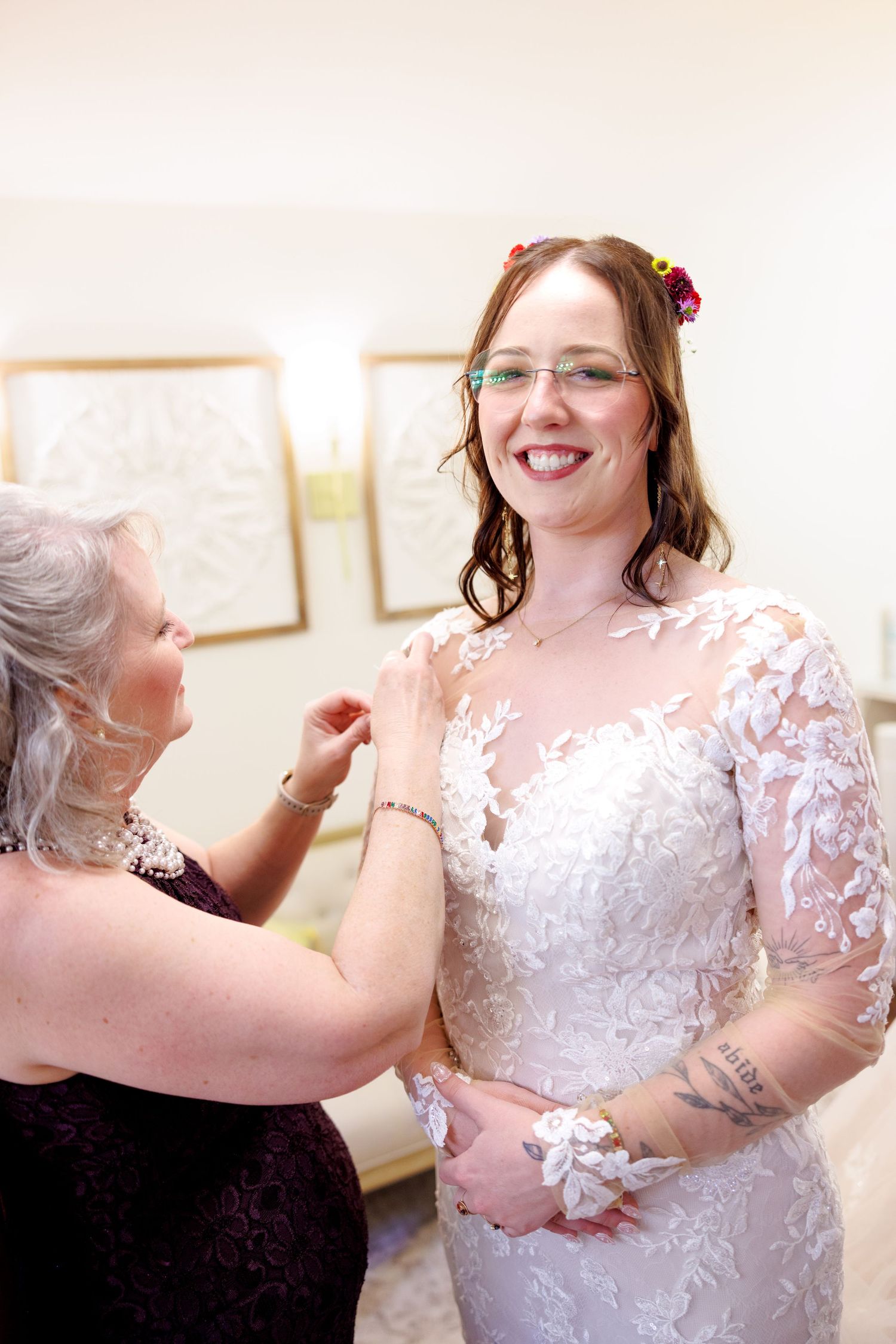 A smiling bride in lace wedding dress with floral hair accessories gets help from an older woman in a burgundy dress.