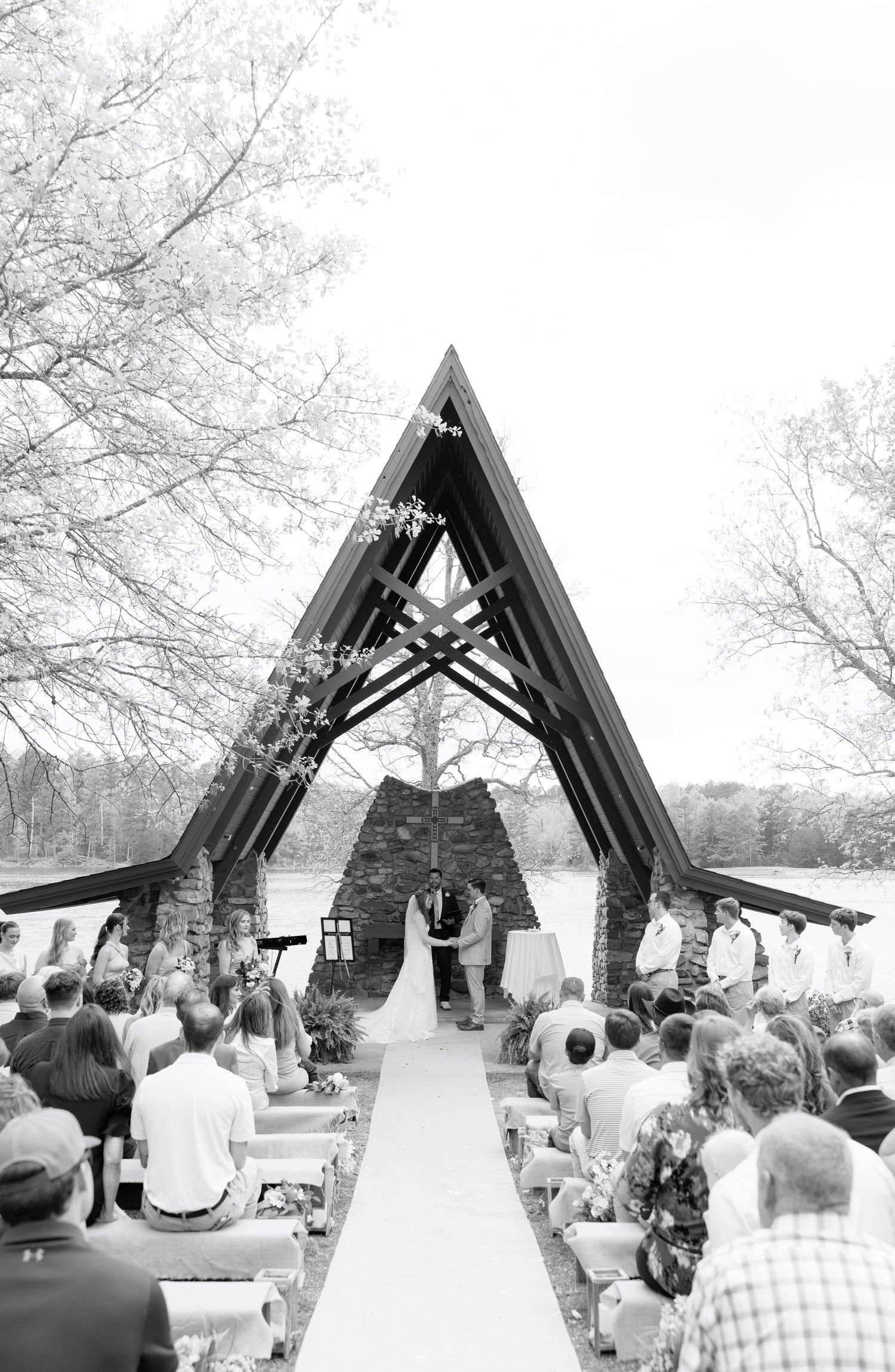 Black and white outdoor wedding ceremony under A-frame arch with stone backdrop, guests seated on white chairs along aisle.