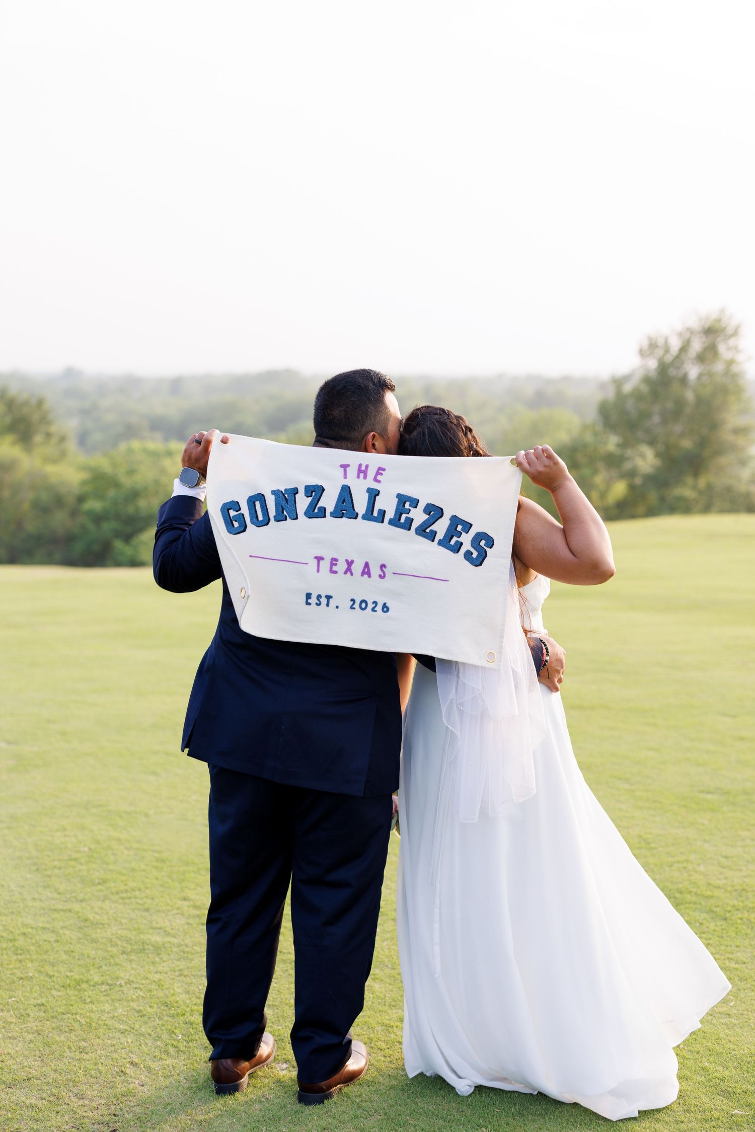 Newlyweds holding a custom banner reading The Gonzalezes Texas Est. 2024 on a golf course.